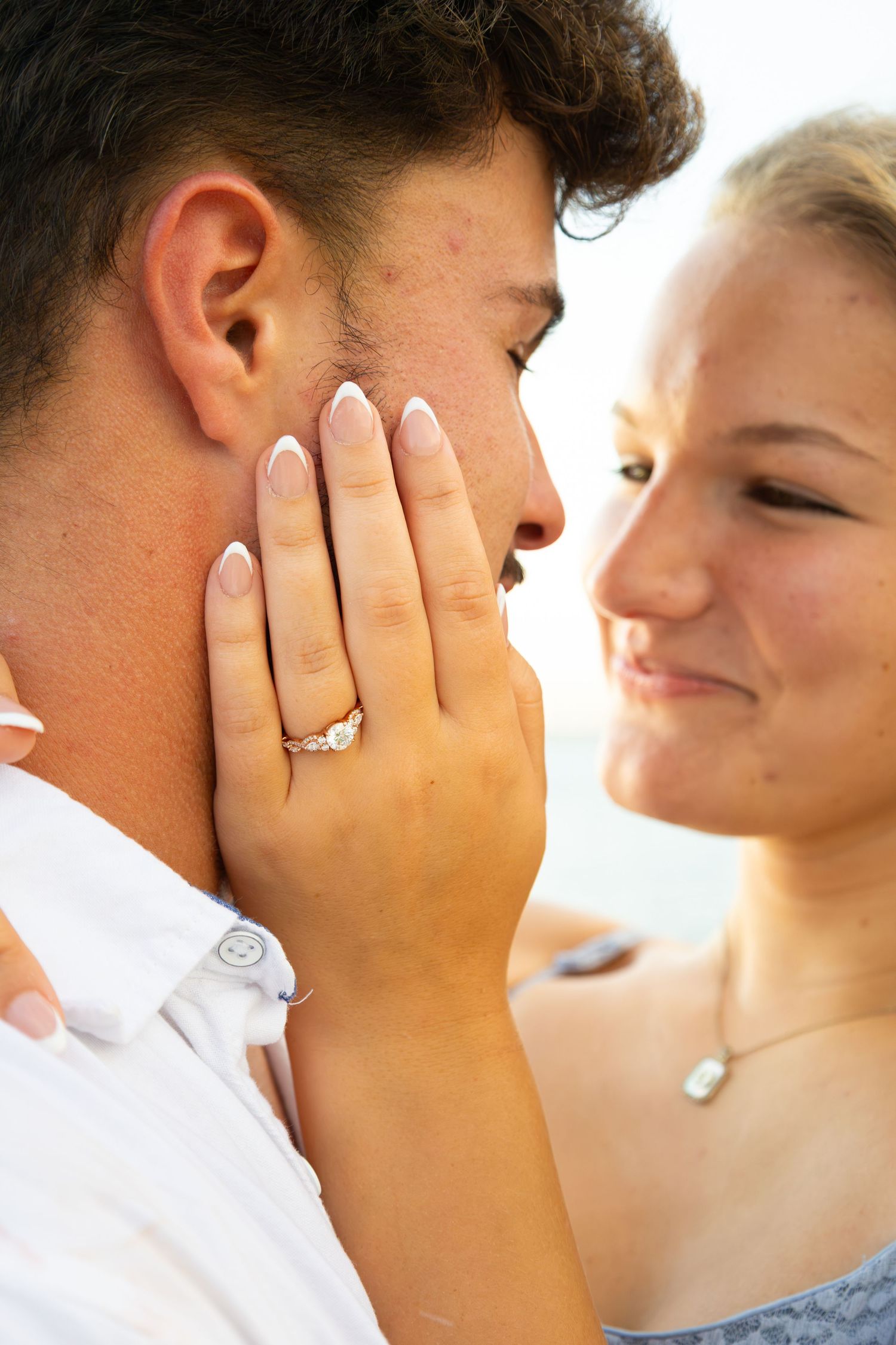 Young couple sharing a romantic moment, woman showing engagement ring while tenderly touching man's face outdoors.
