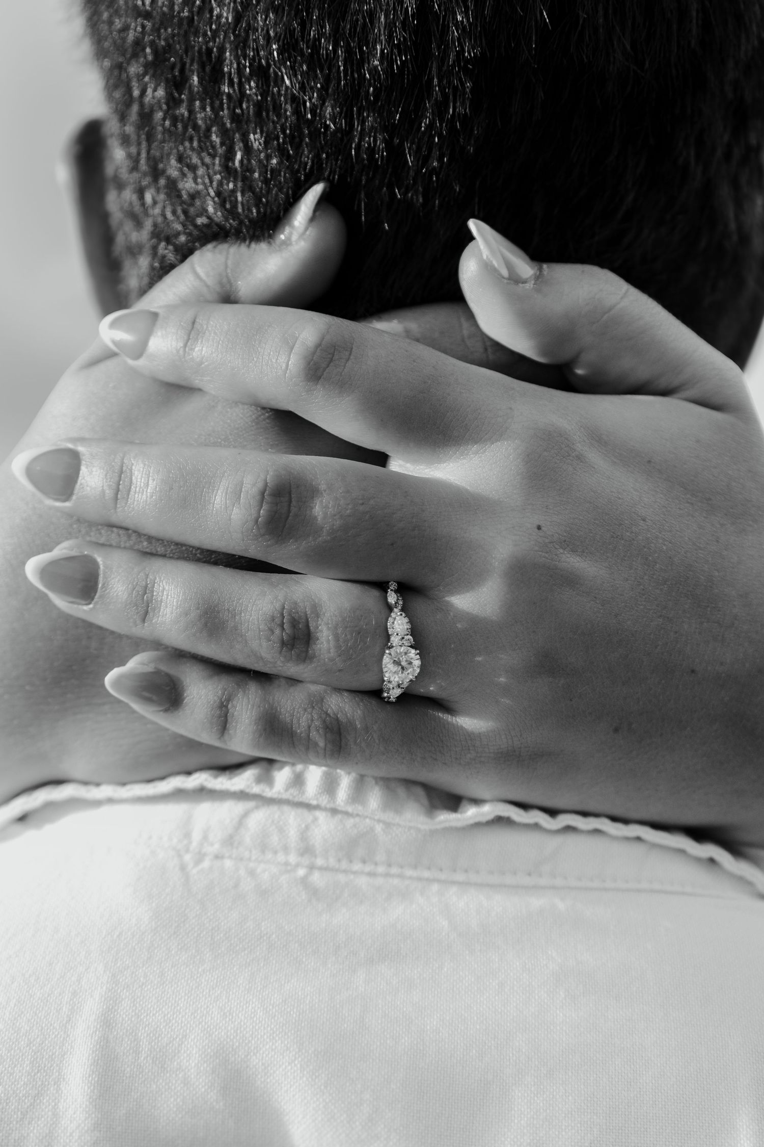 Close-up black and white photo of a woman's hand with a diamond engagement ring resting on a man's neck.