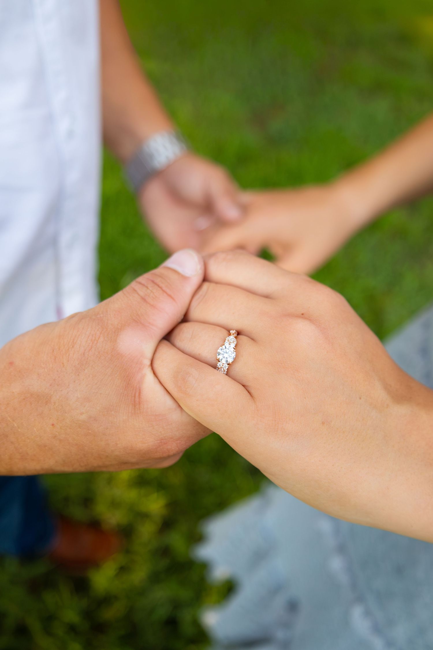 Close-up of couple holding hands outdoors, woman wearing a sparkling three-stone diamond engagement ring on green background.