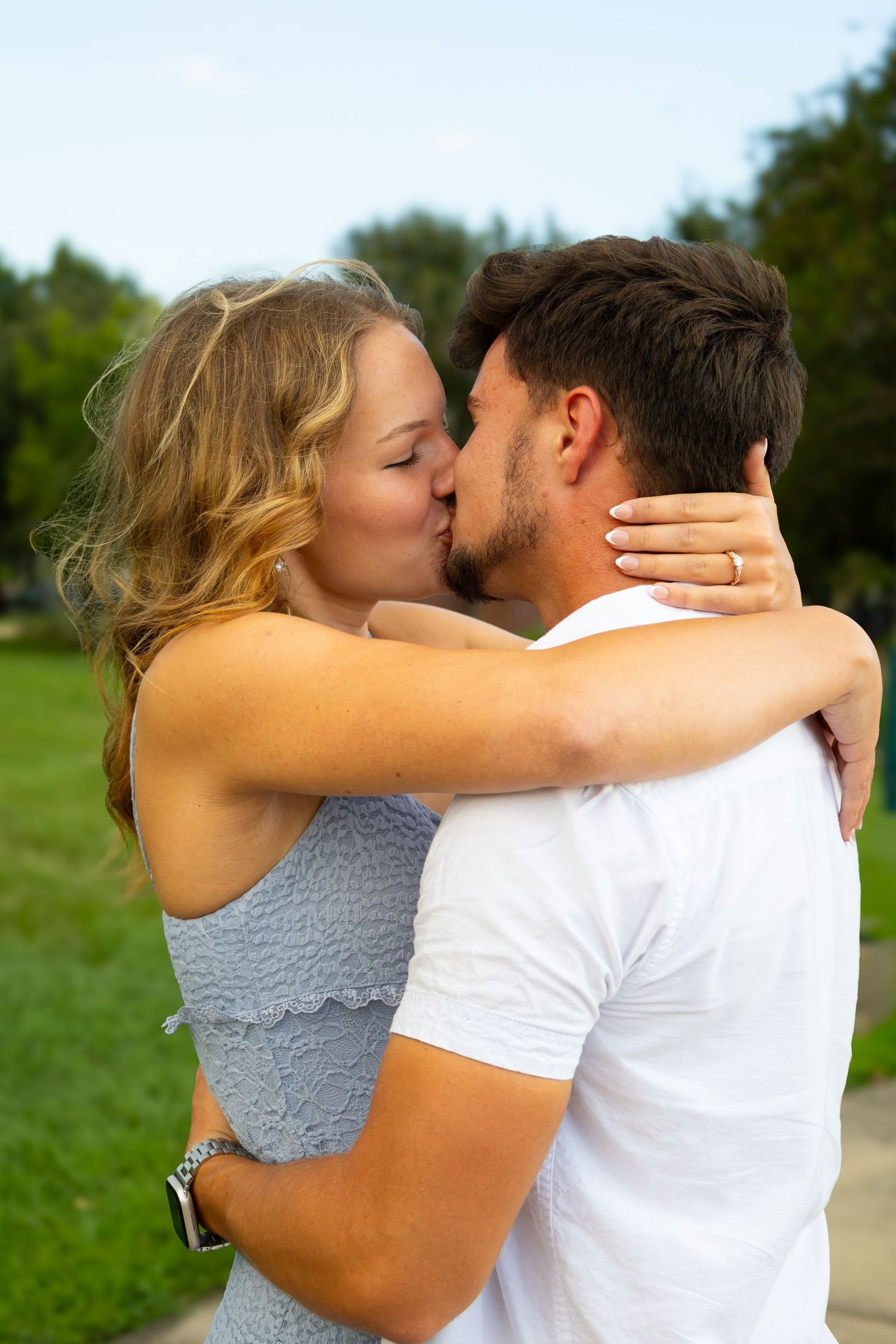 A young couple sharing a romantic kiss outdoors in a park, woman in blue dress embracing man in white t-shirt.