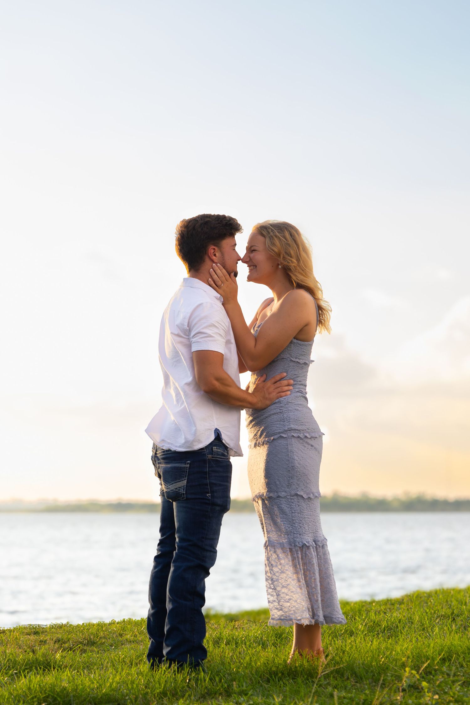 Romantic couple embracing and laughing together on green grass by a lake at sunset.