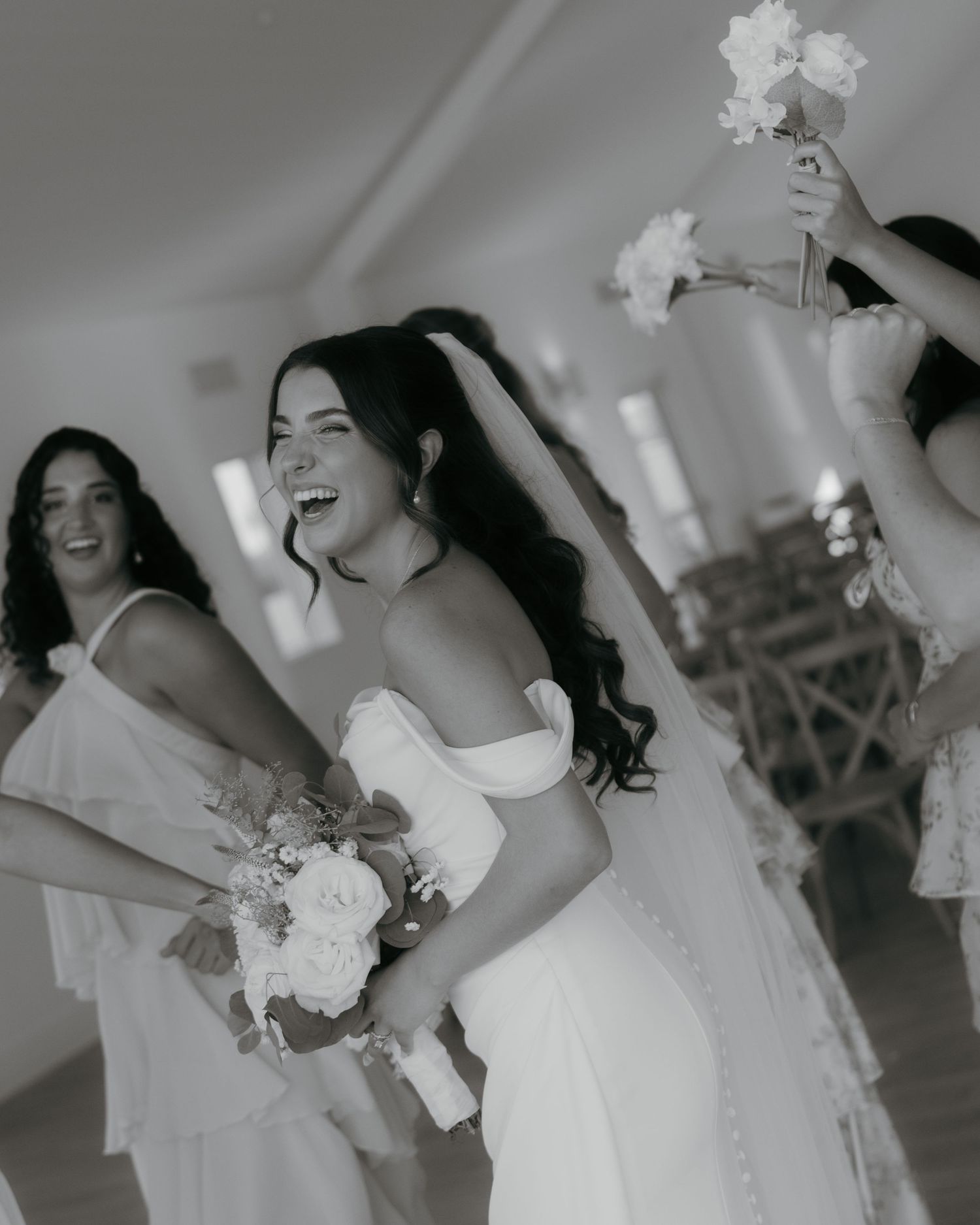 Bride laughing joyfully with bridesmaids holding bouquets in black and white wedding photo.