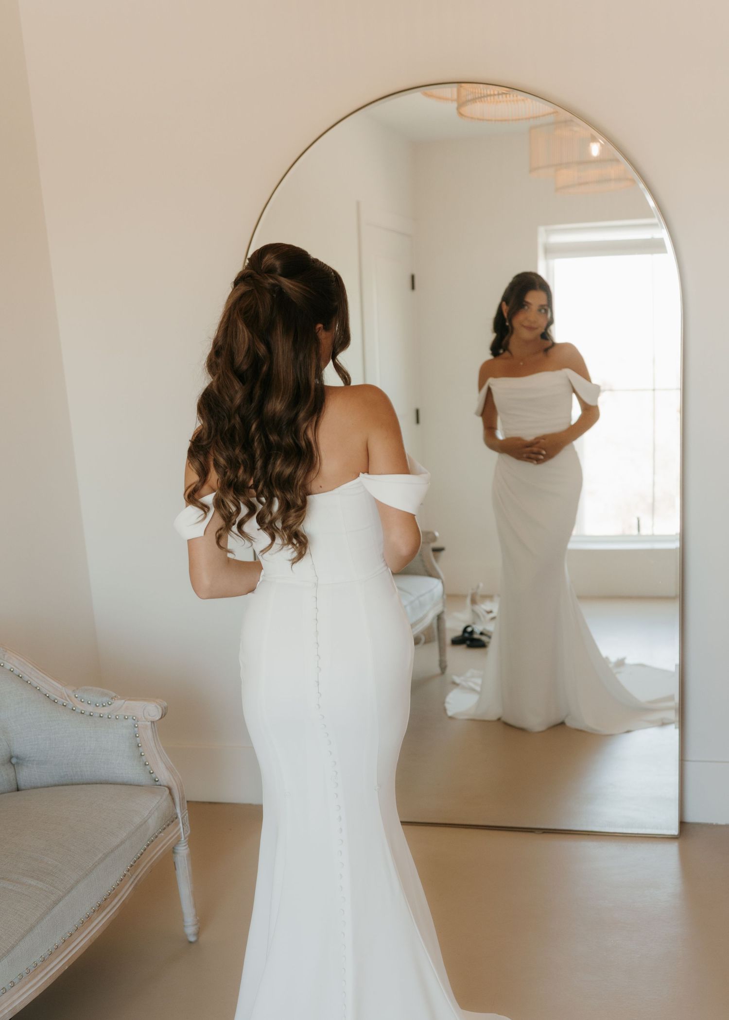 Bride in off-shoulder white mermaid wedding gown admiring her reflection in a large arched mirror.