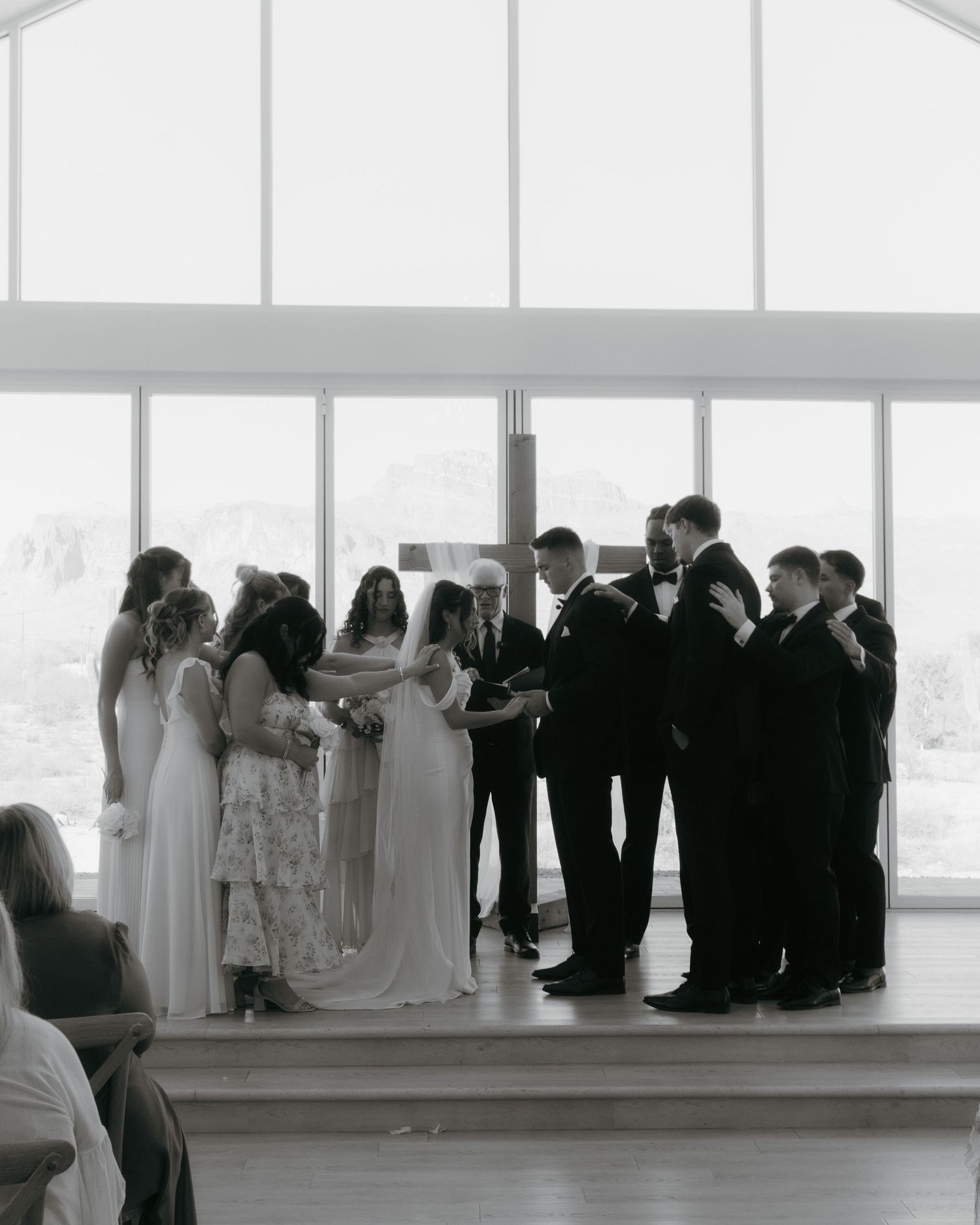 Black and white wedding ceremony photo with bridal party gathered around couple exchanging vows before large windows.