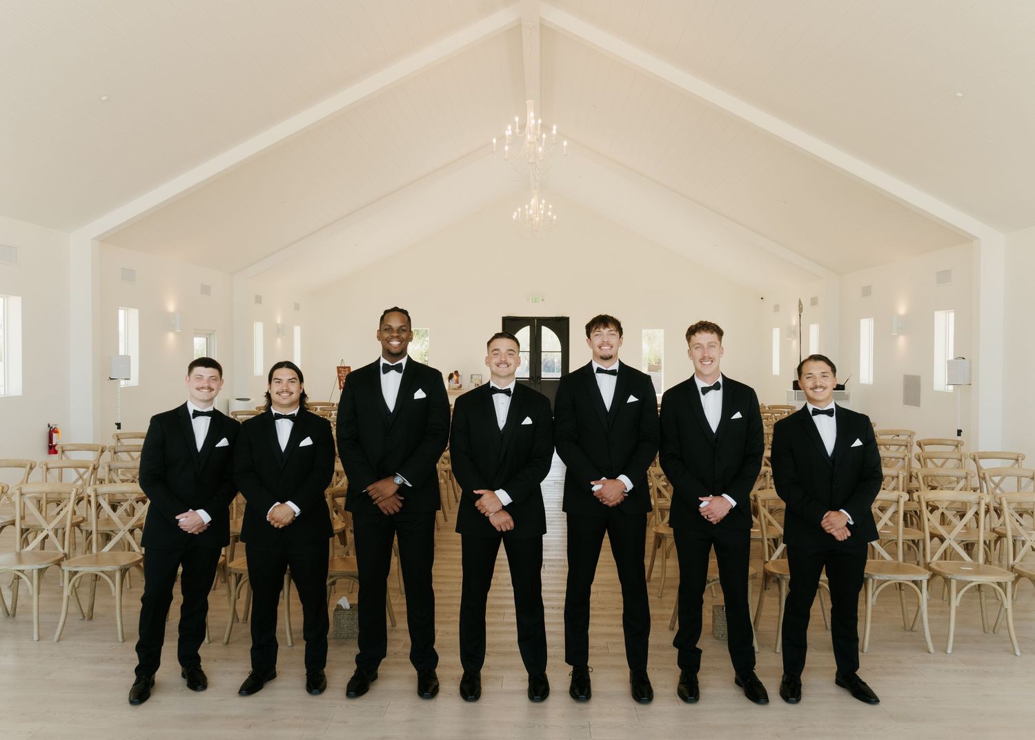 Seven groomsmen in black tuxedos with bow ties stand in a bright white wedding chapel with vaulted ceilings.