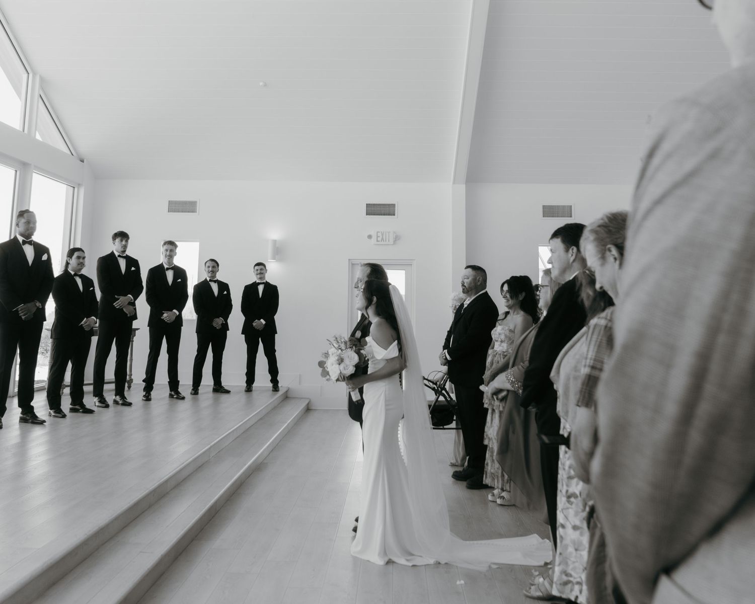 Black and white wedding ceremony photo with bride holding bouquet facing groomsmen lined up against a bright white wall.