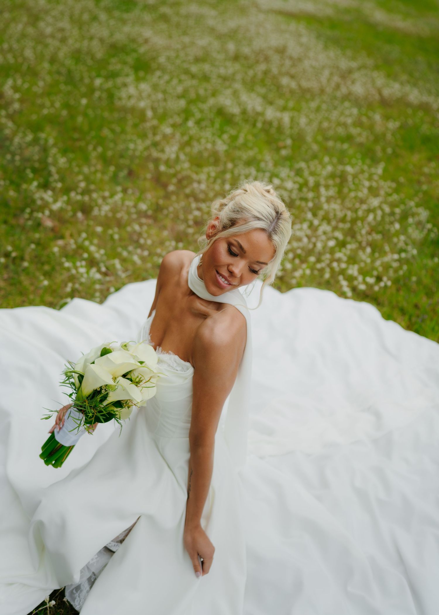 Elegant blonde bride in white halter wedding dress sits on grass holding white floral bouquet outdoors.