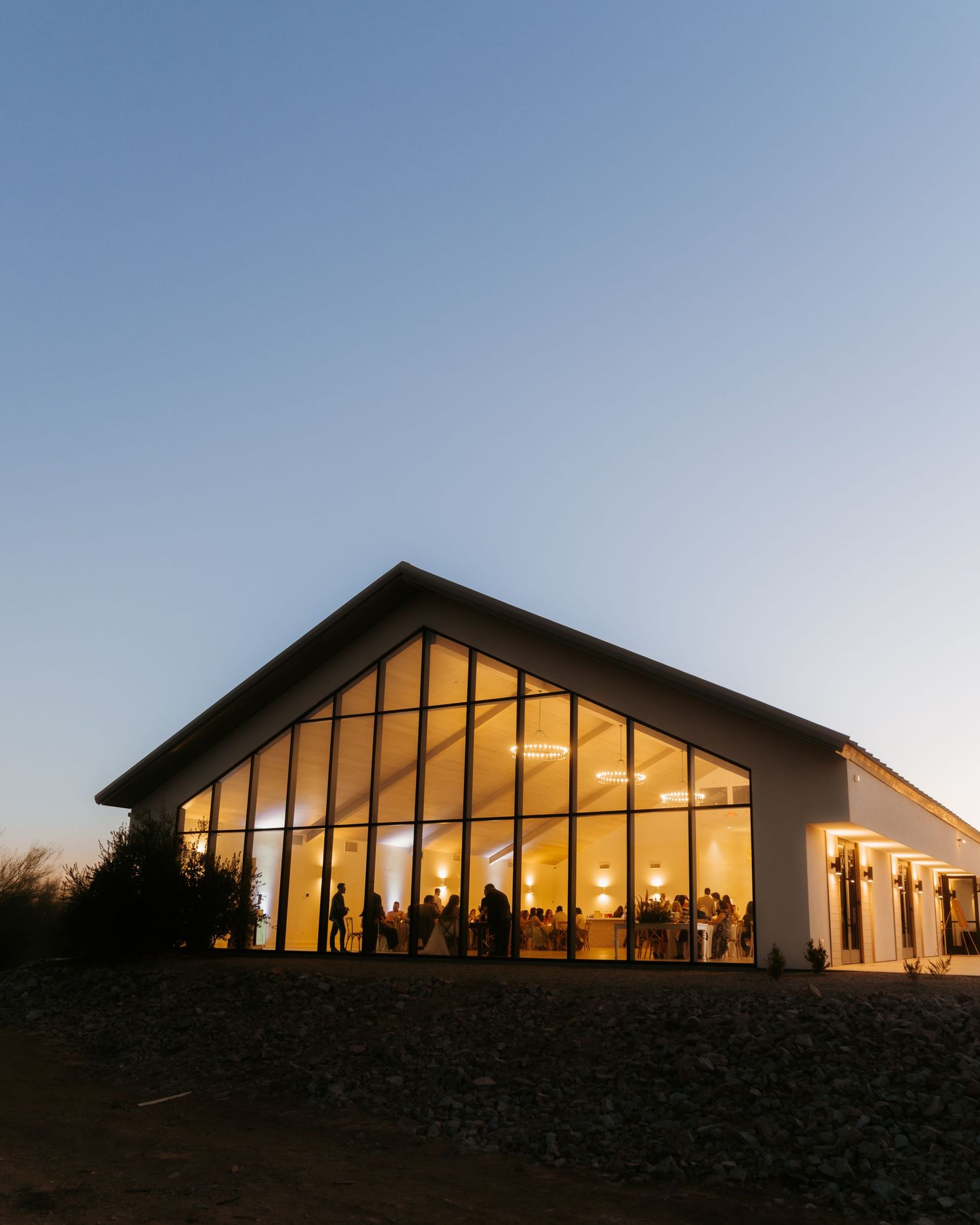 Elegant event venue with floor-to-ceiling windows glowing warmly at dusk, guests visible inside during an evening celebration.