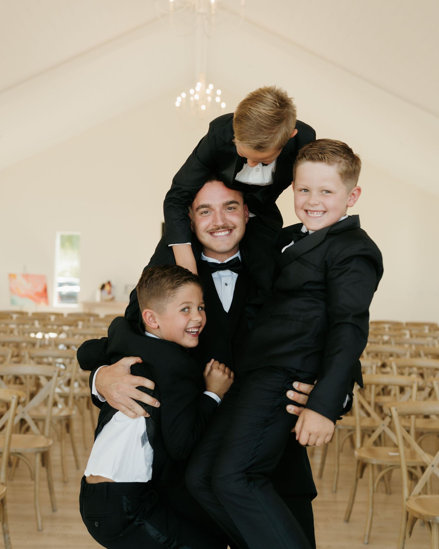 Groom in black tuxedo joyfully embracing three young boys in matching suits at wedding venue.
