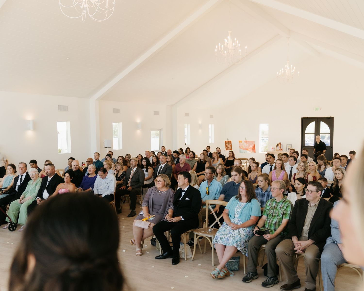 Guests seated in rows of wooden chairs inside a bright white chapel during a wedding ceremony.