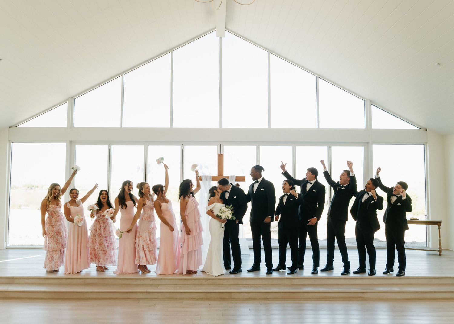 Wedding party celebrating in bright venue, bridesmaids in pink gowns and groomsmen in black suits cheering with arms raised.
