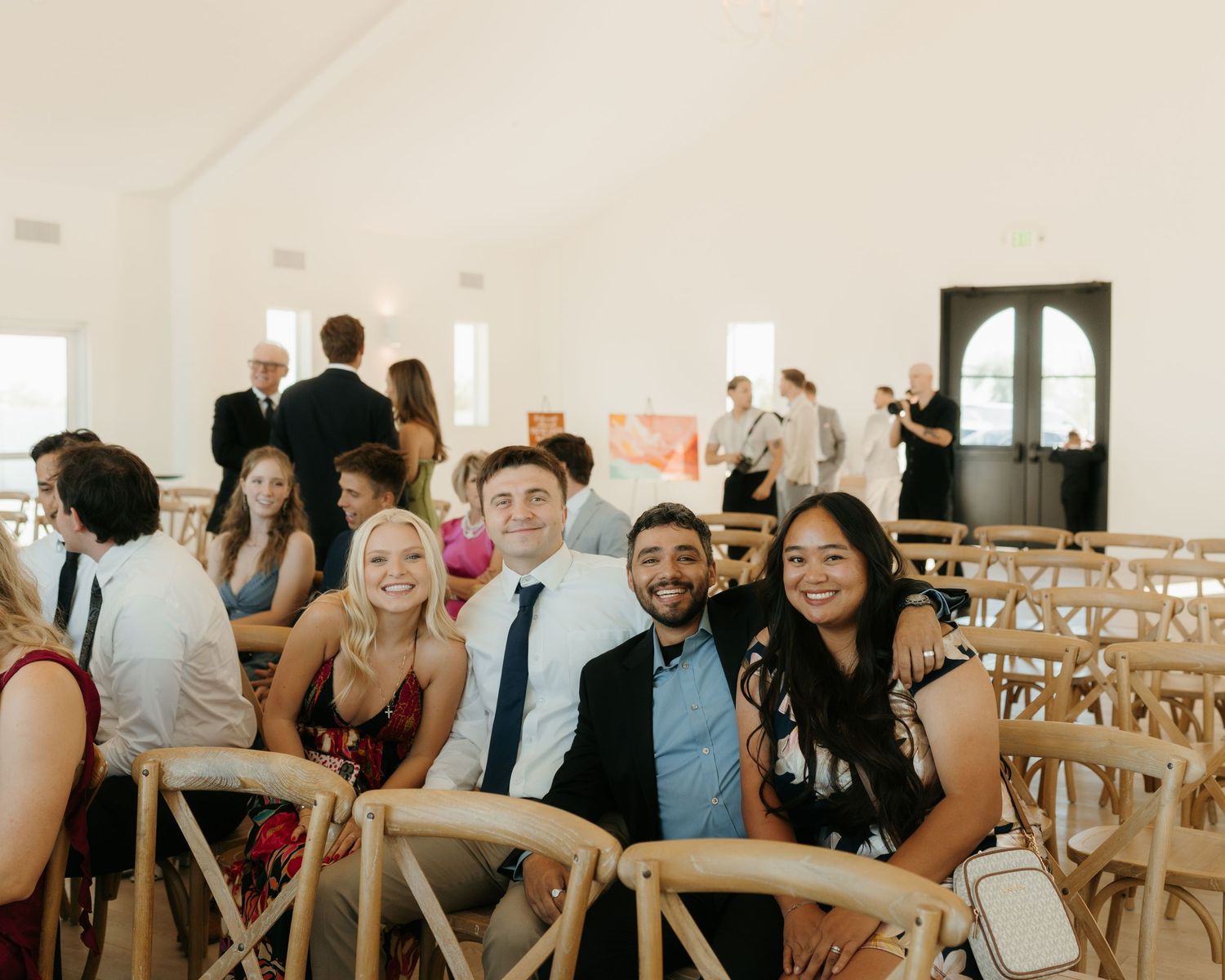 Guests smiling at a wedding ceremony seated in wooden chairs inside a bright white venue.