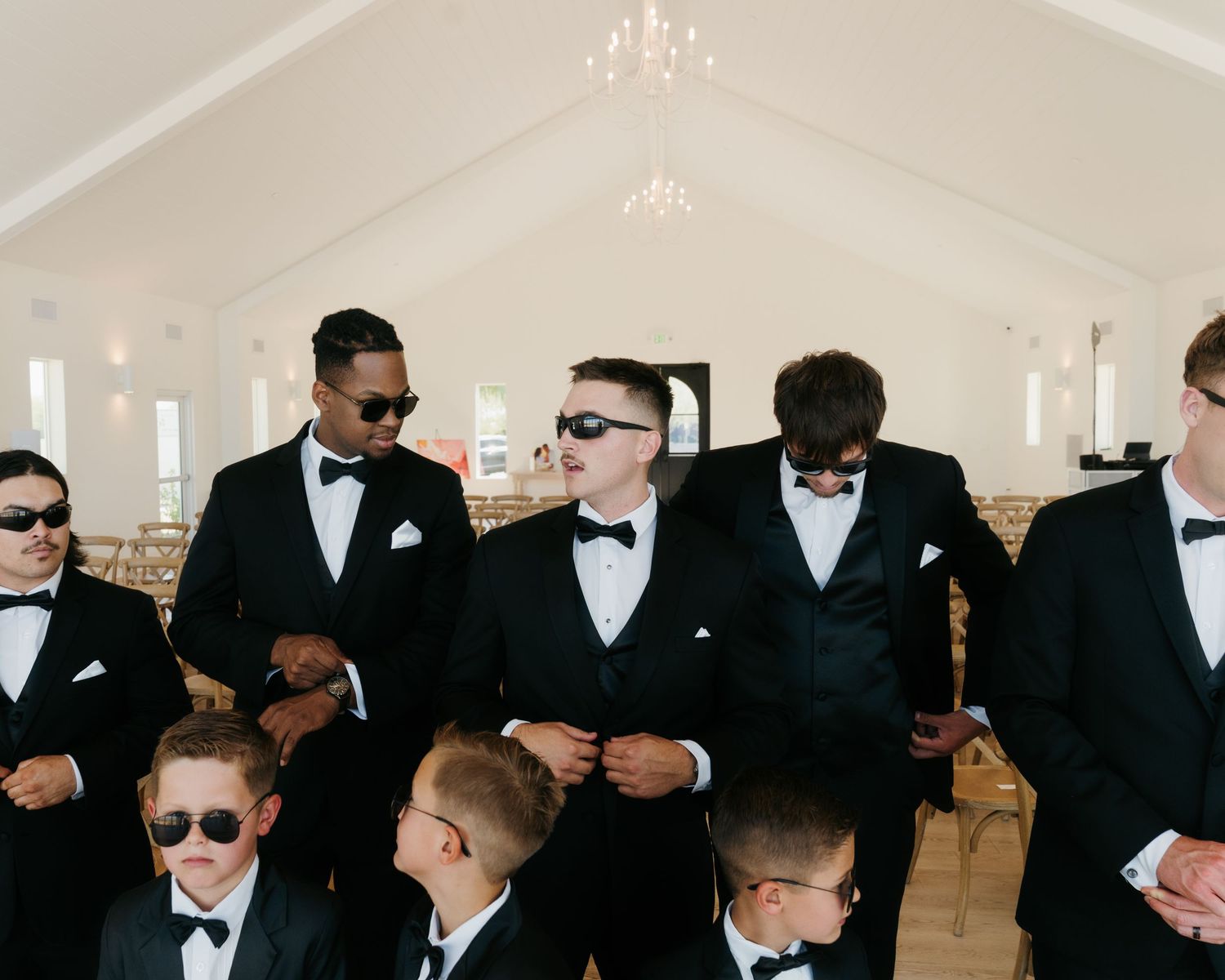 Groom and groomsmen in black tuxedos and sunglasses posing inside elegant wedding venue.
