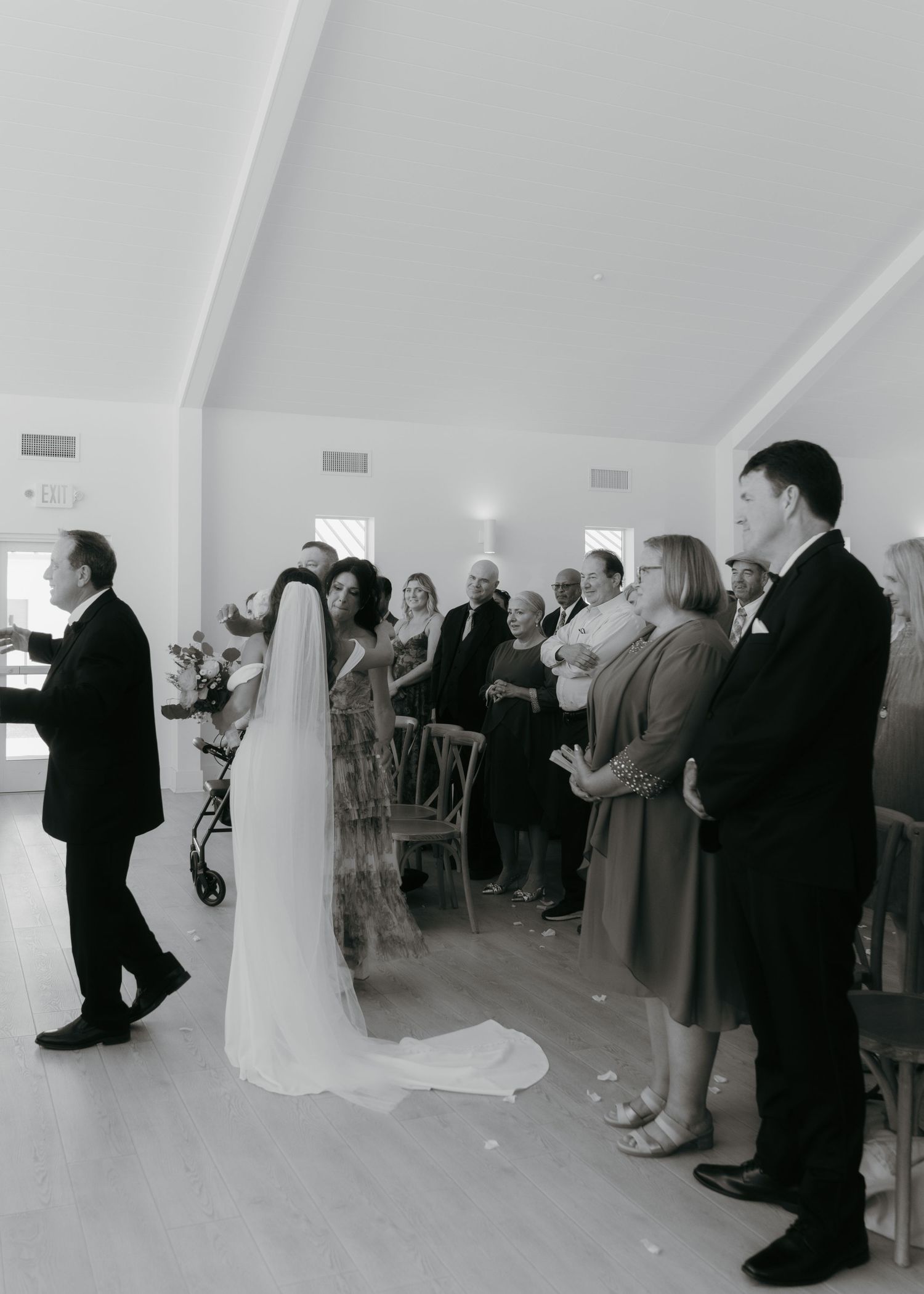 Black and white photo of a bride walking down the aisle at an indoor wedding ceremony with guests standing.