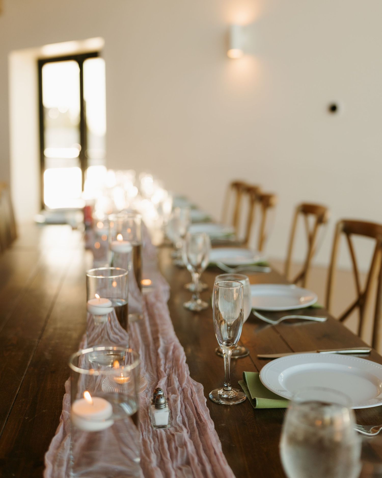 Elegant wedding reception table set with candles, wine glasses, and blush cheesecloth runner in warm light.
