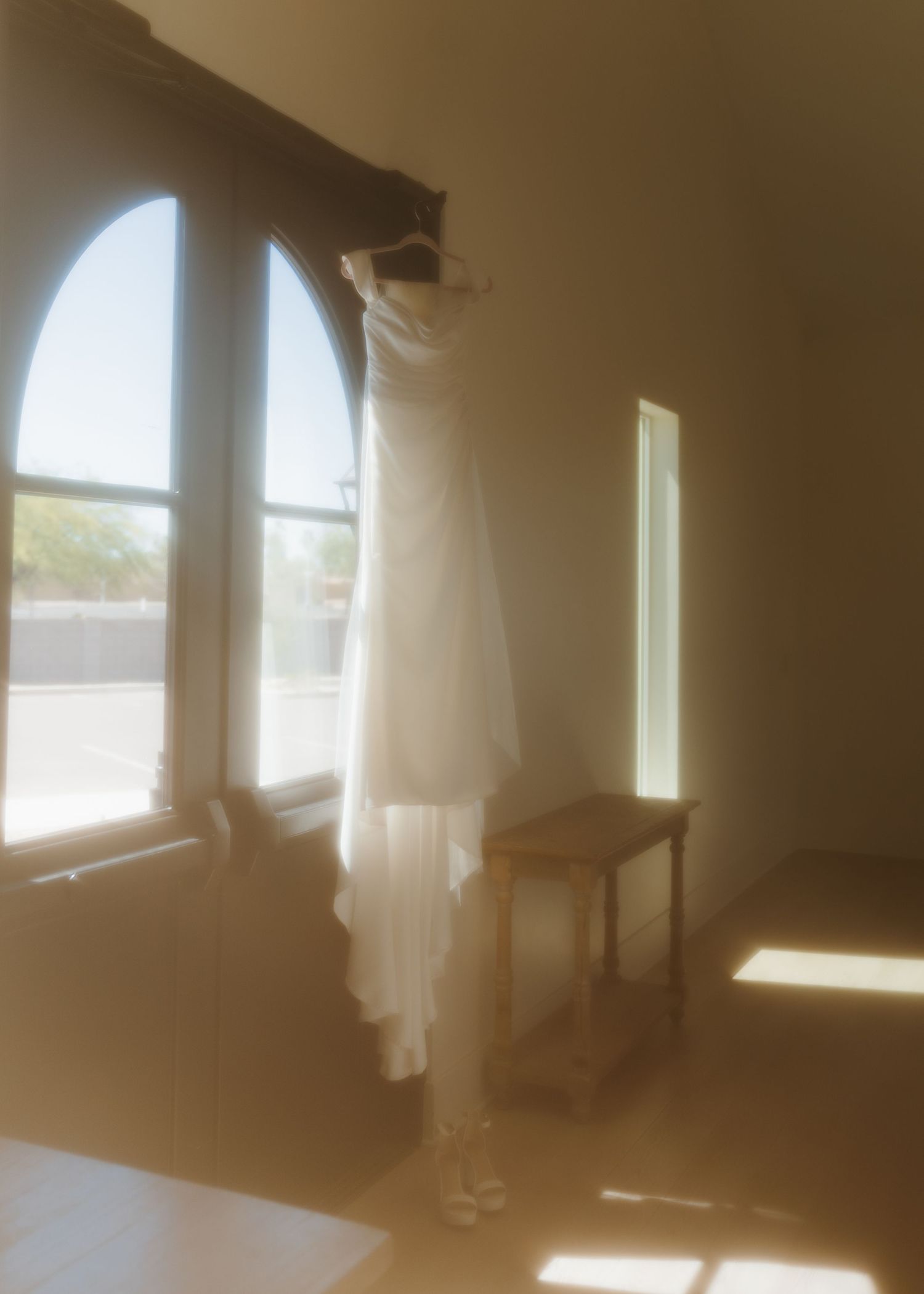 White wedding dress hanging on an arched window in a sunlit bridal suite room with wooden side table.