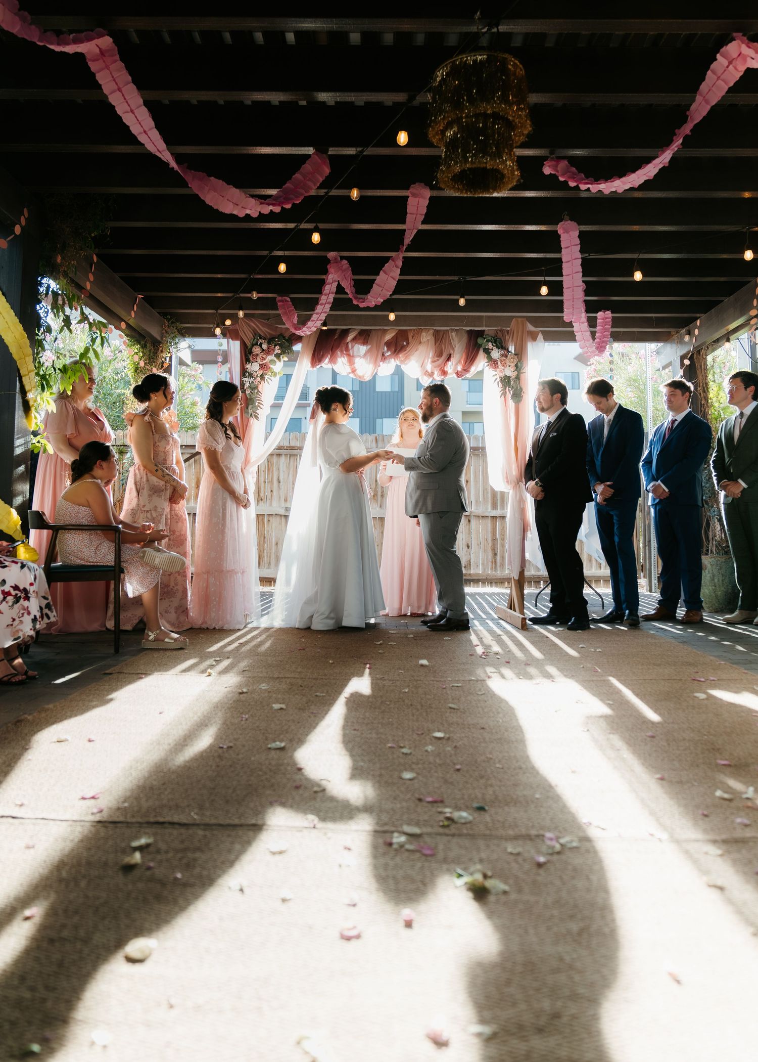 Bride and groom exchange vows at a decorated indoor ceremony with pink streamers and string lights.