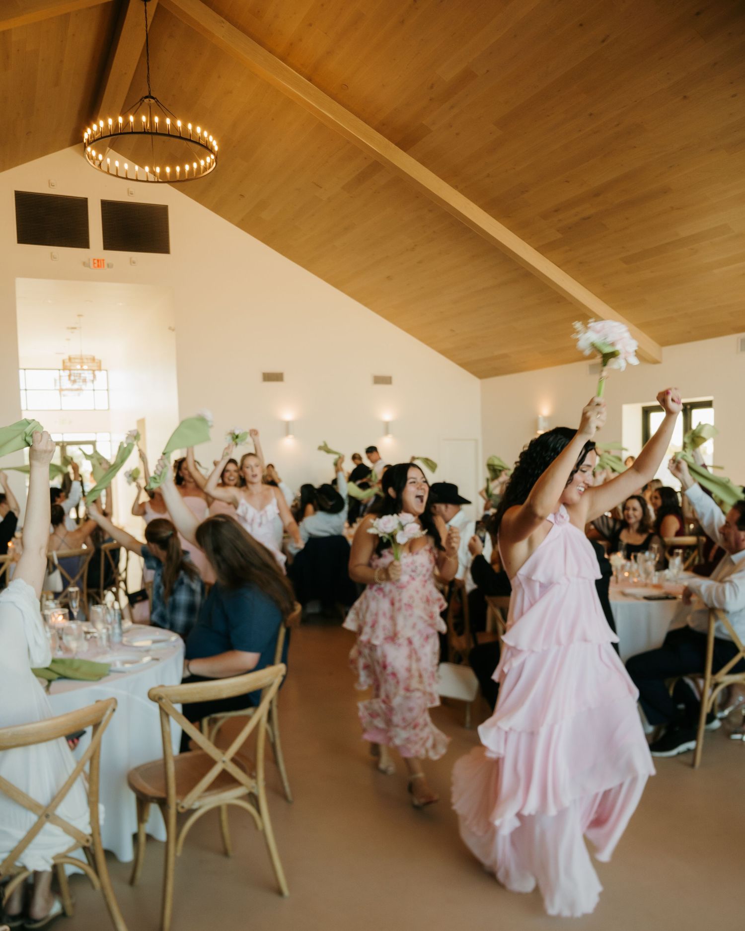 Guests raise flowers and palm leaves in celebration at an elegant wedding reception in a white venue.