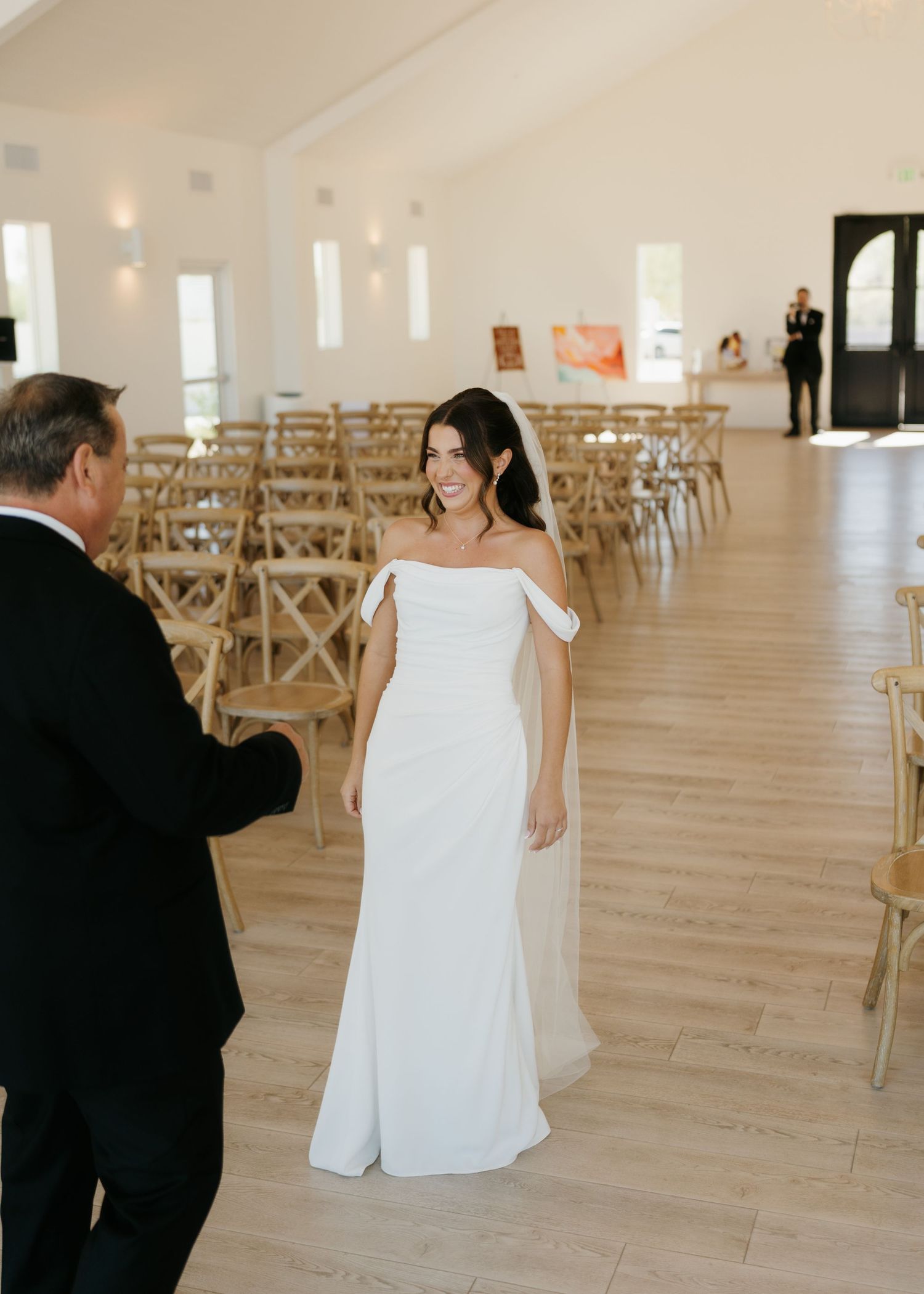 Bride in off-shoulder white gown smiling during first look ceremony in elegant white venue with wooden chairs.