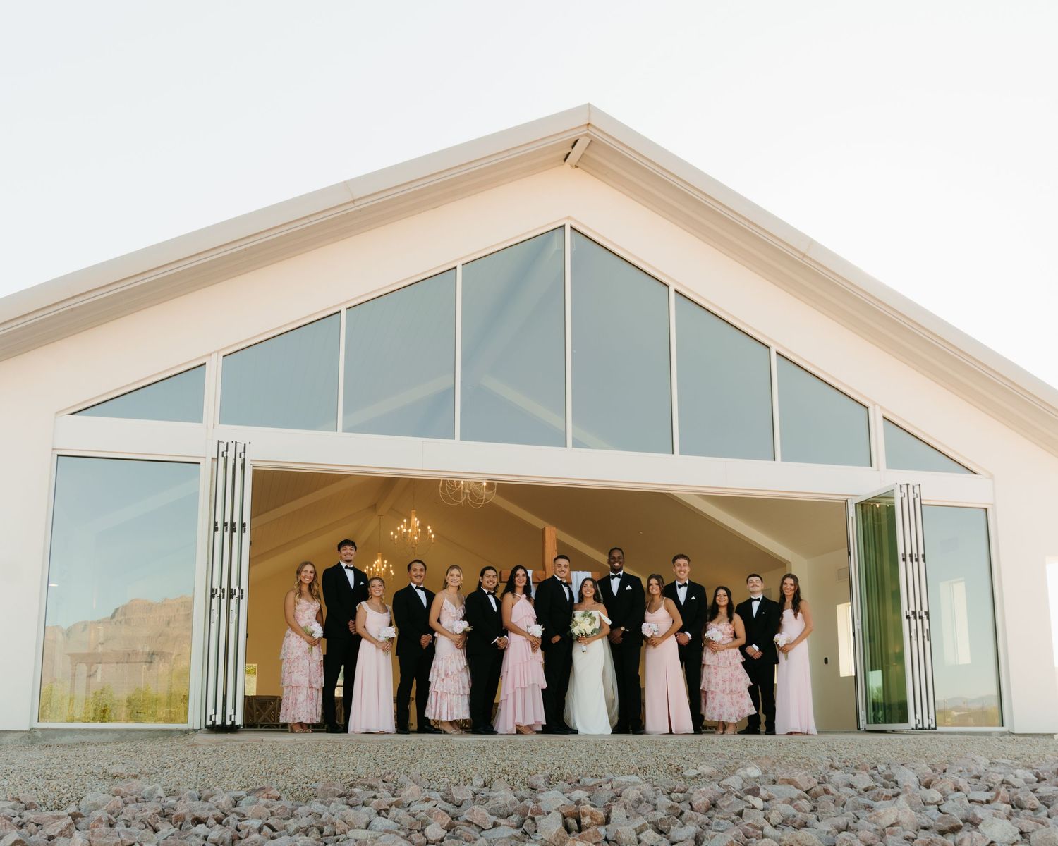 Wedding party poses in front of a modern white barn venue with large glass windows at sunset.
