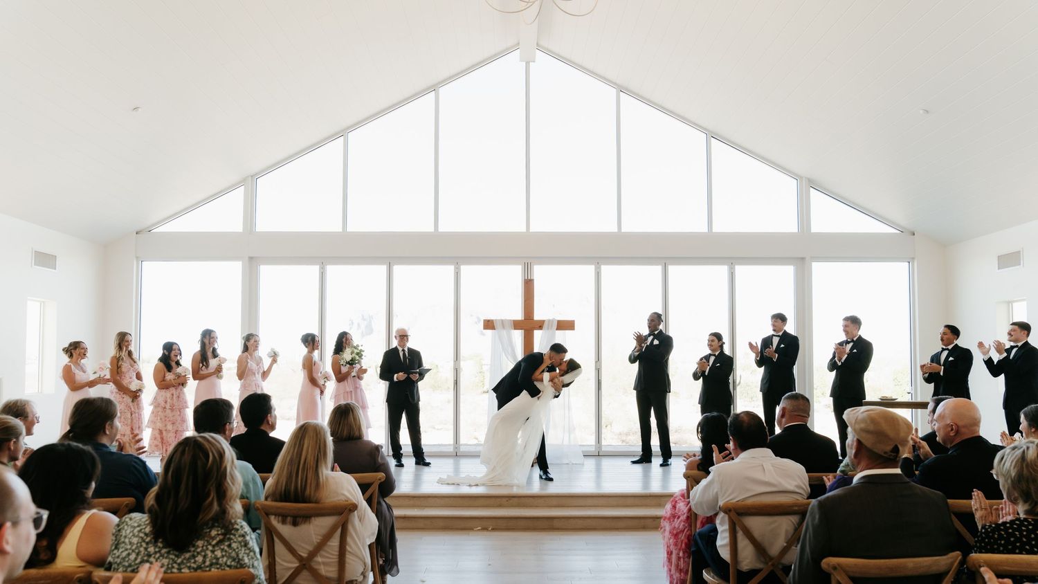 Bride and groom share first kiss at elegant chapel wedding ceremony with bridesmaids and groomsmen applauding.