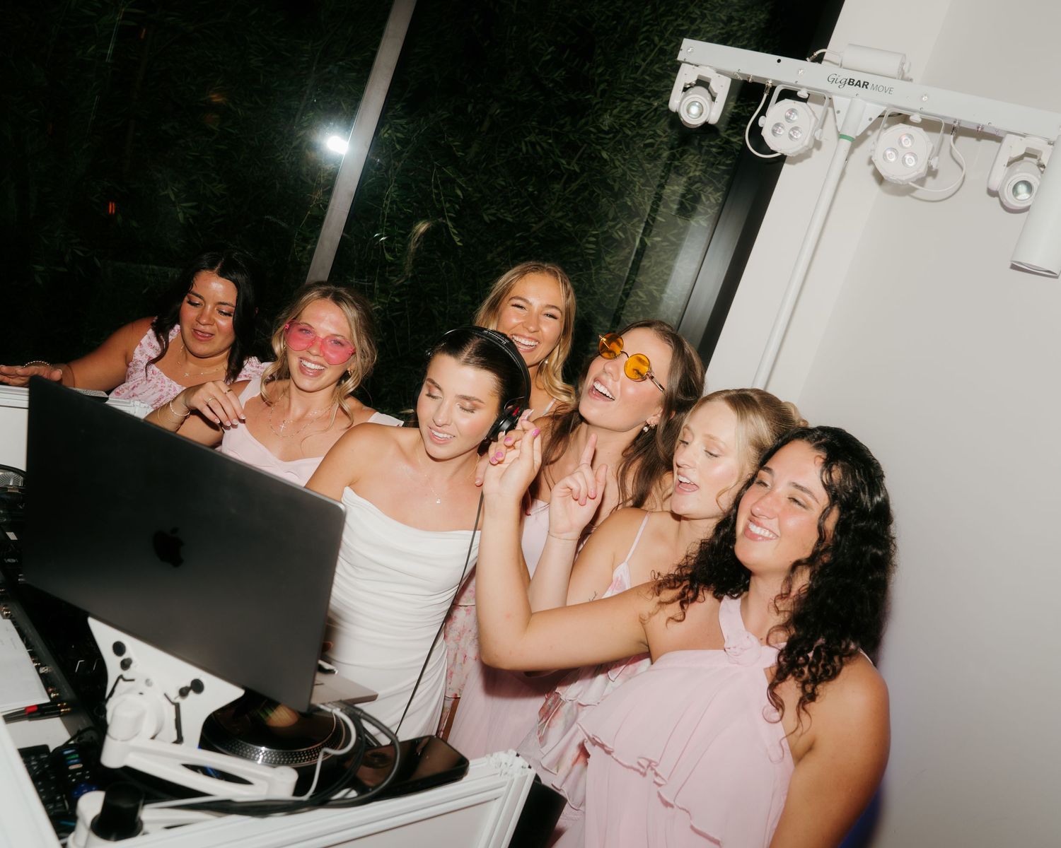Bride and bridesmaids in pink dresses pose together laughing at the DJ booth during a lively wedding reception.