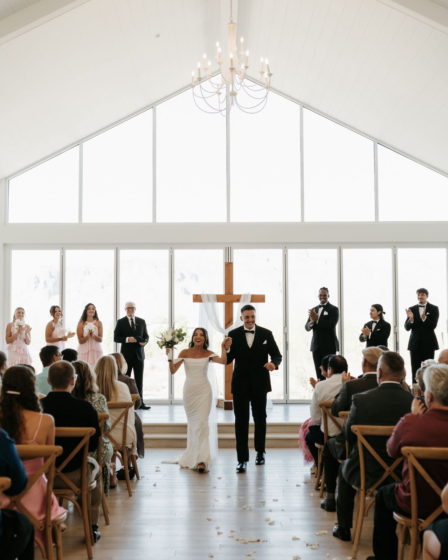 Bride and groom walk down the aisle in a bright chapel with a wooden cross, surrounded by wedding party and guests.