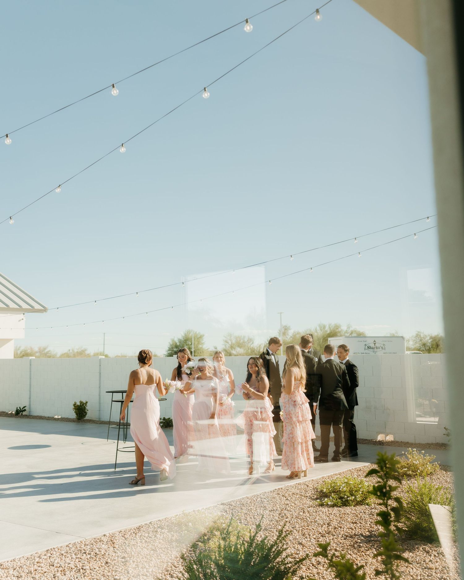 Bridesmaids in pink dresses gather outdoors at a wedding reception under string lights in a sunny courtyard.