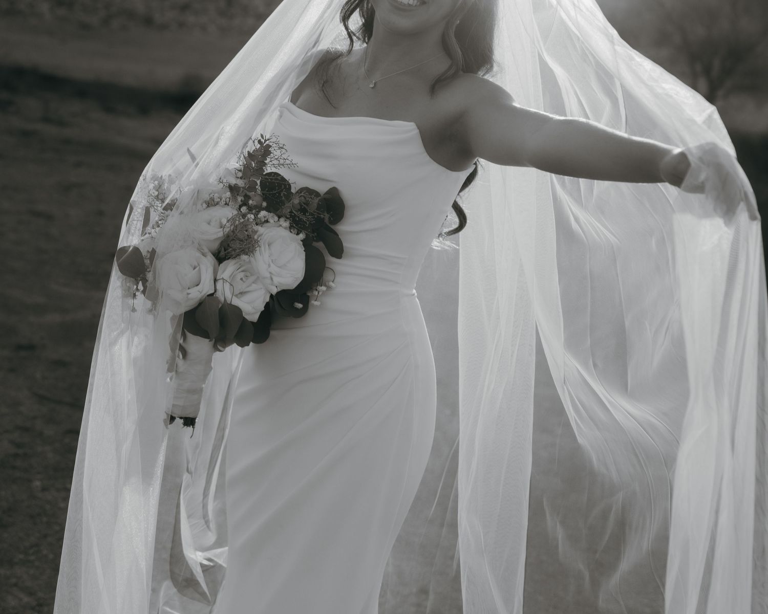 Black and white photo of a bride in a white gown holding a bouquet, her veil flowing gracefully in the wind.