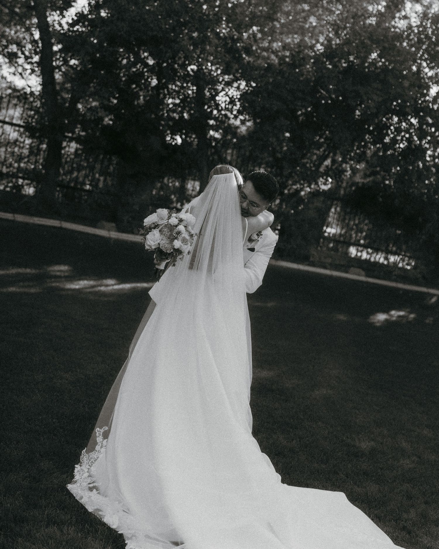 Black and white photo of bride in cathedral veil and gown holding bouquet in garden setting.