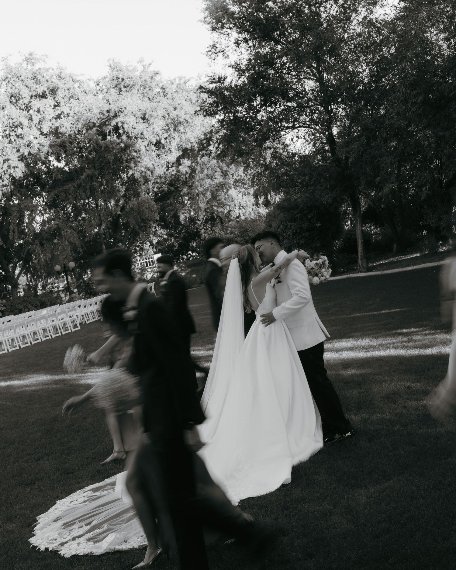 Black and white motion blur photo of bride and groom kissing in a garden surrounded by guests.