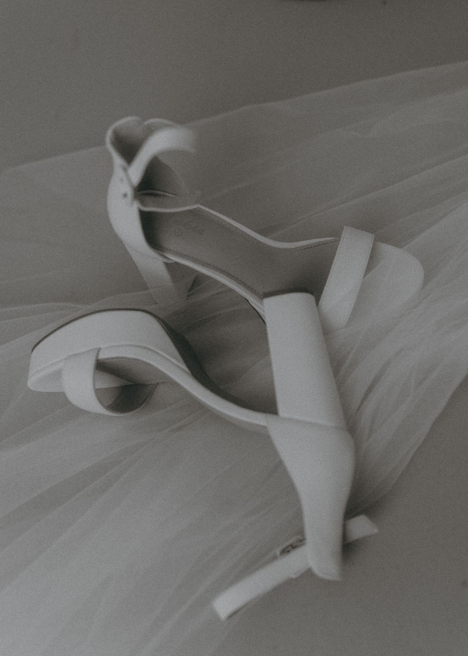 White bridal heeled sandals with ankle strap resting on soft tulle fabric in elegant monochrome wedding photography.