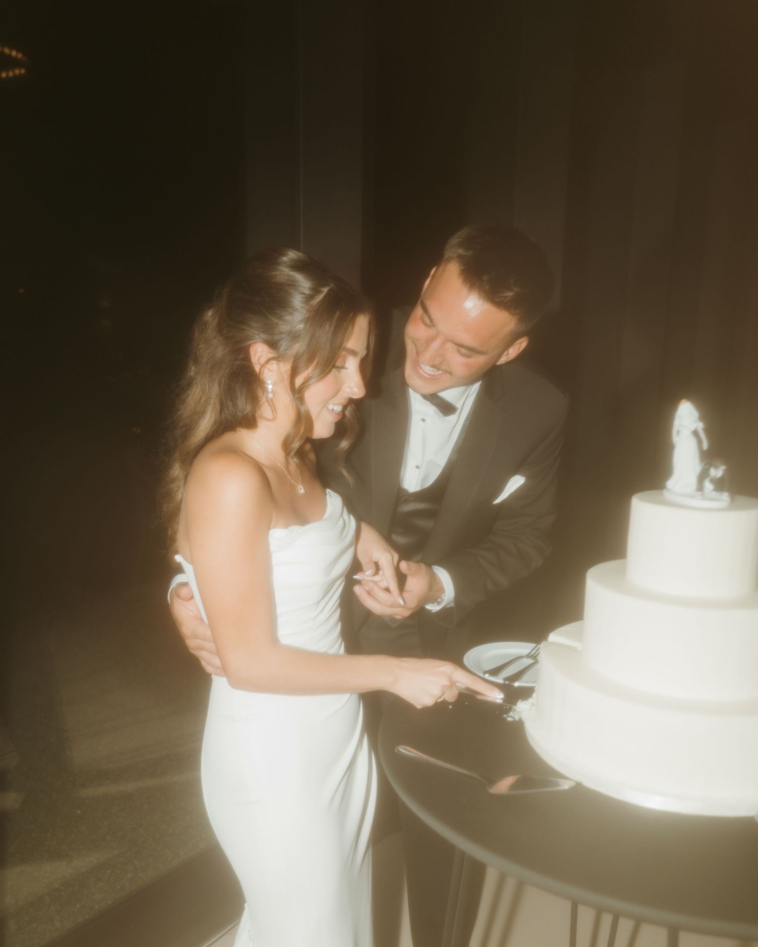 Smiling bride and groom in formal attire cut their white three-tier wedding cake together at their reception.