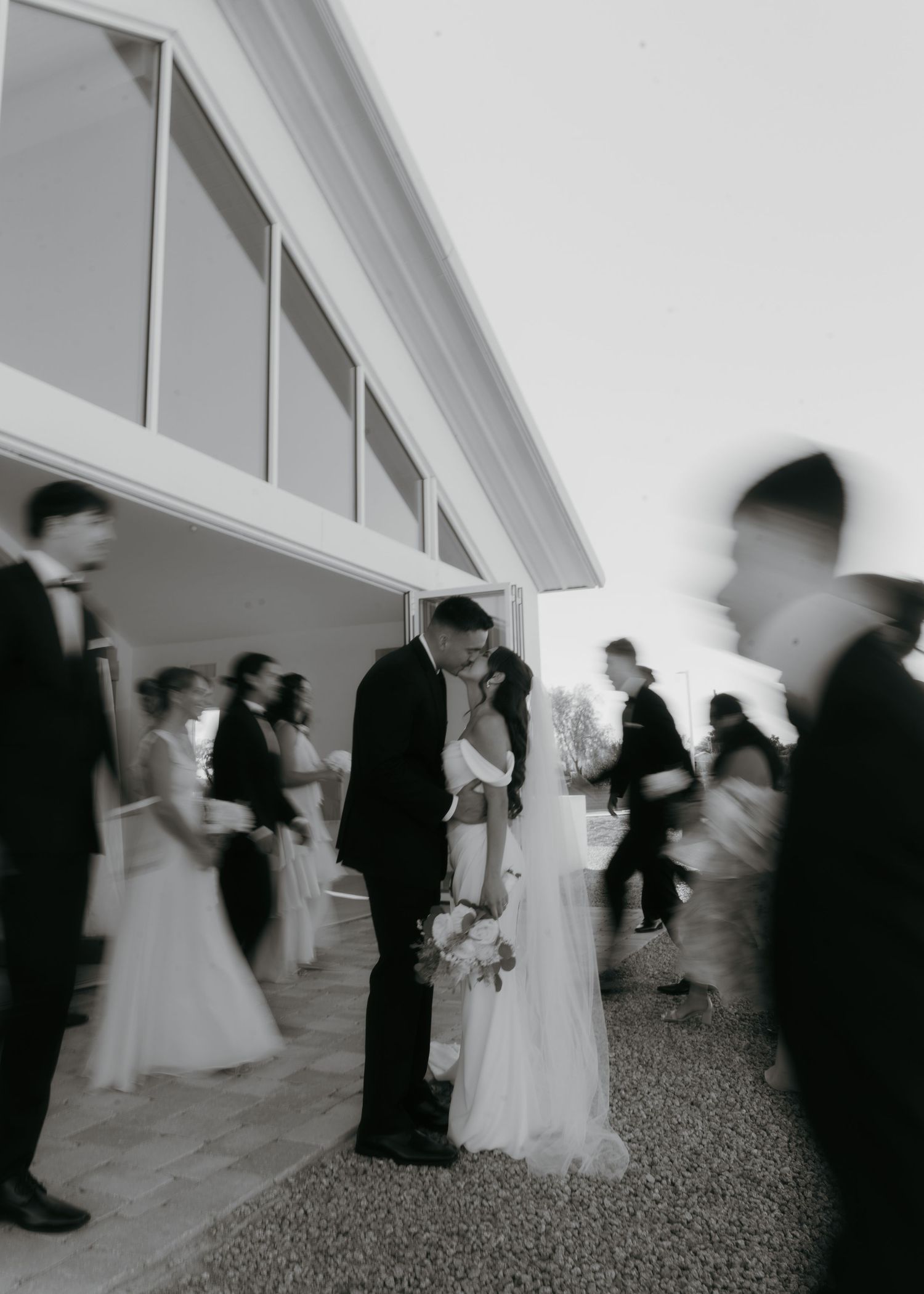 Black and white wedding photo of bride and groom sharing a kiss outdoors with guests mingling in background.