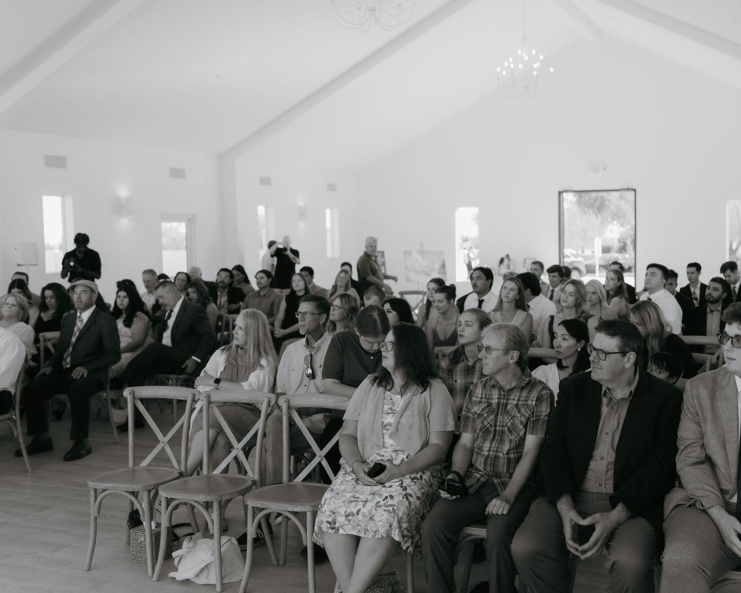 Black and white photo of guests seated in rows at an indoor wedding ceremony in a bright white venue.