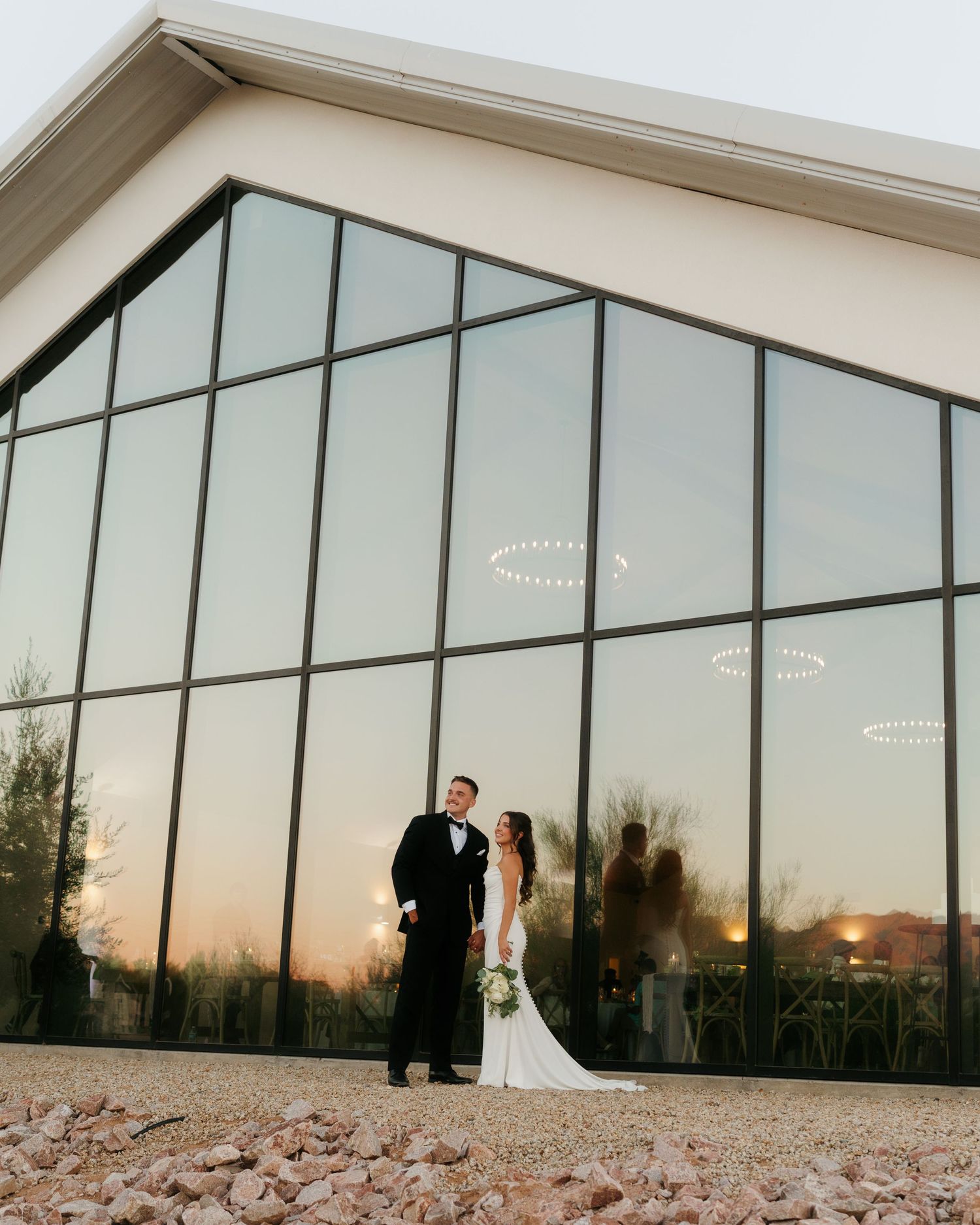 Bride and groom posing at sunset before large glass wall of modern wedding venue.