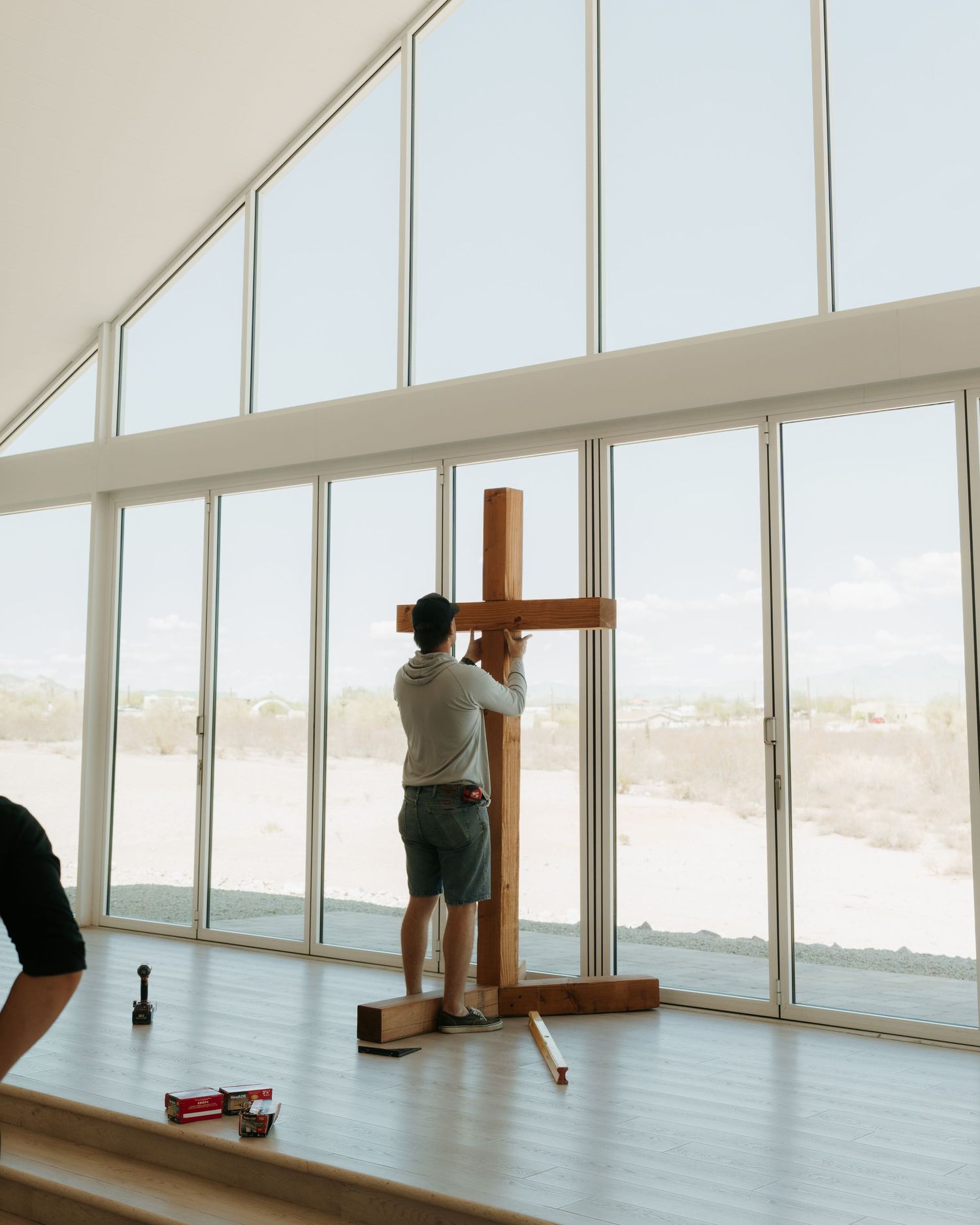 Man assembling a wooden cross in a modern church with large floor-to-ceiling windows and desert landscape views.
