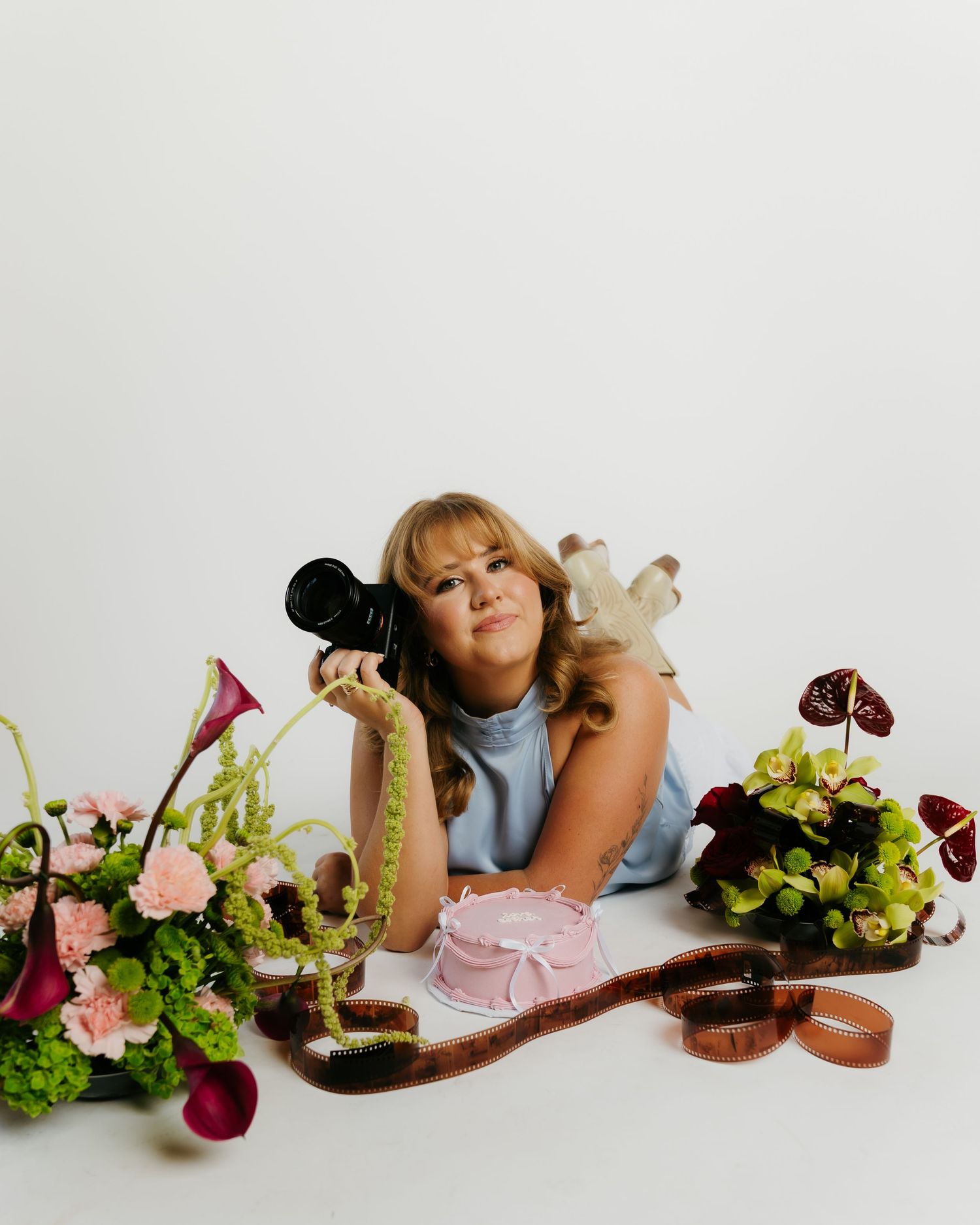 Woman lying among colorful floral arrangements, holding a camera, with film strips and a snake nearby.