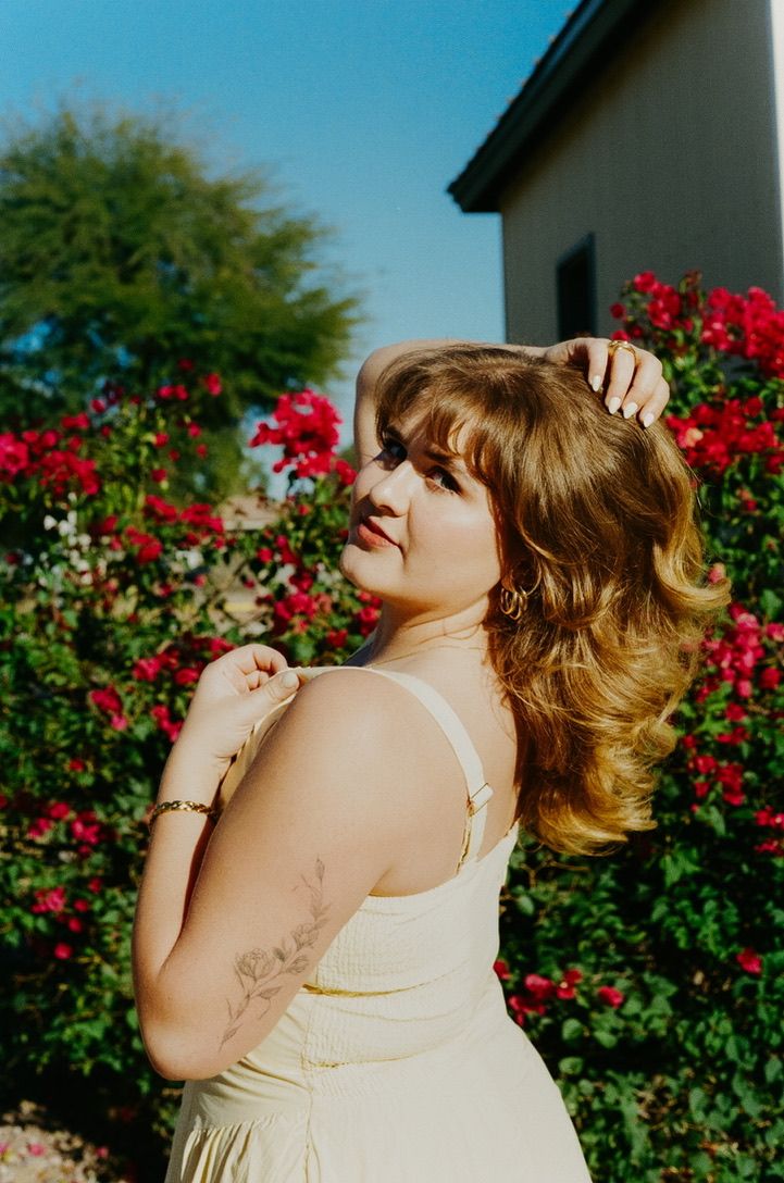 Young woman in yellow sundress posing among vibrant red roses in a sunny outdoor garden setting.
