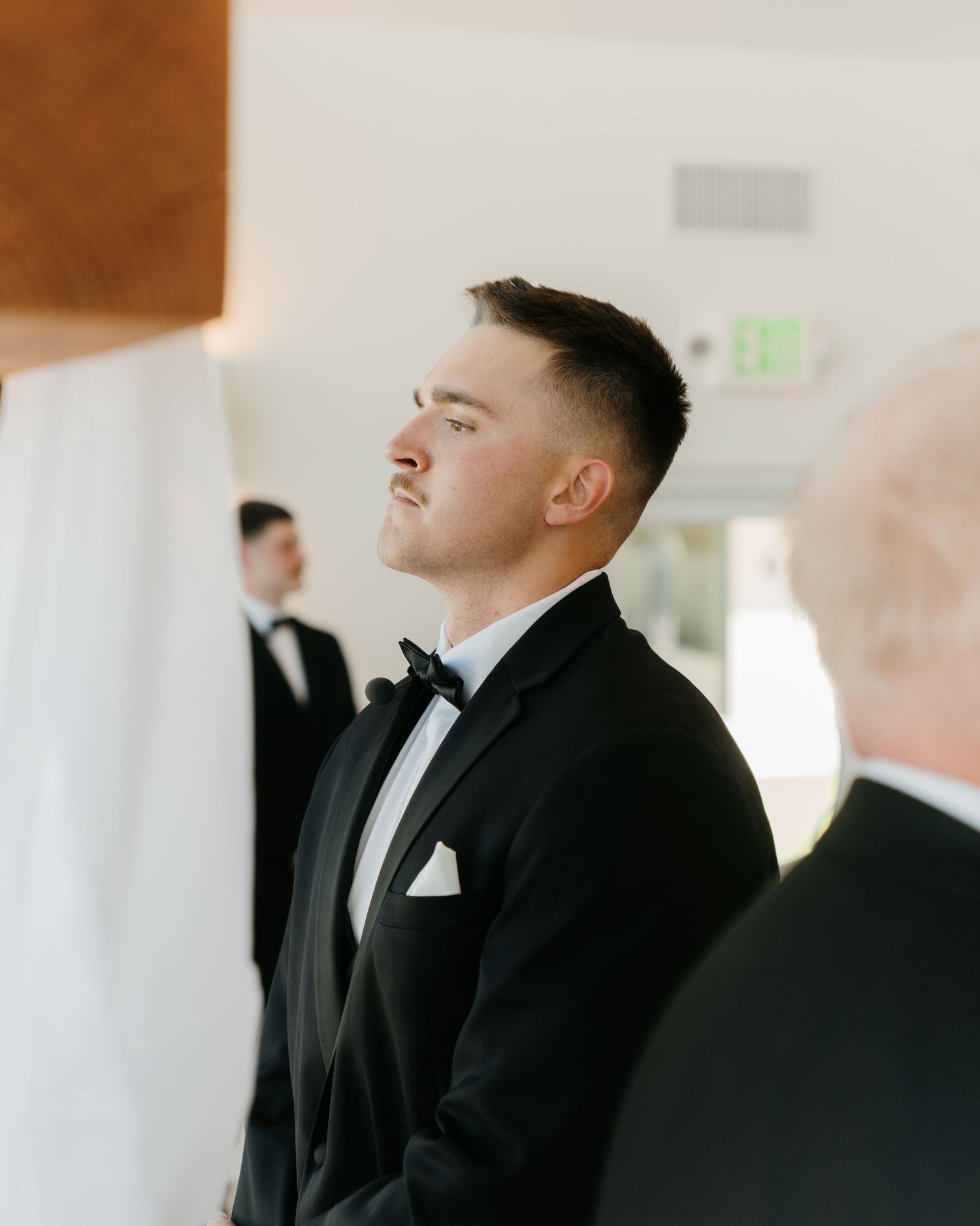 Groom in black tuxedo and bow tie standing at altar with eyes closed during wedding ceremony.