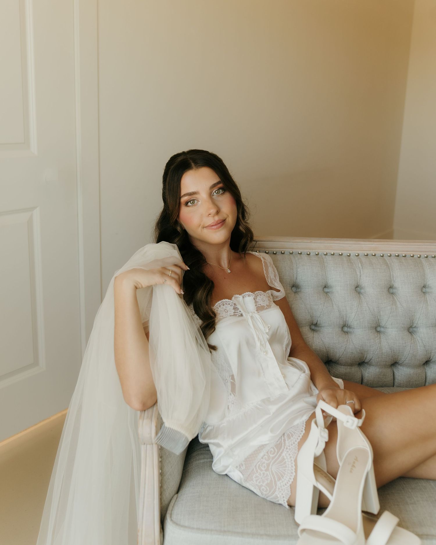 Bride in white lace slip dress and sheer robe sits gracefully on a gray velvet sofa holding bridal heels.