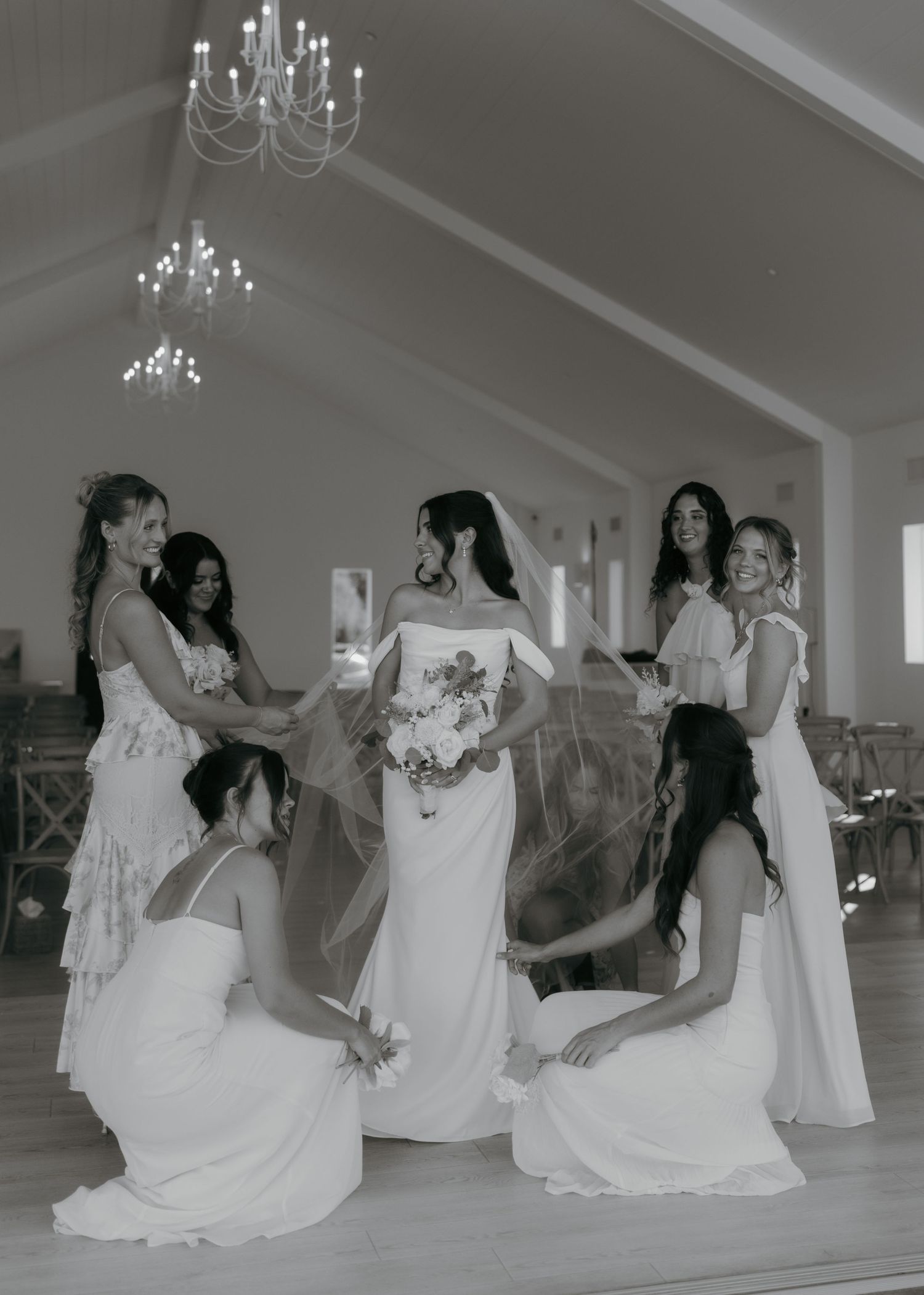 Black and white photo of a bride in a white gown holding bouquet surrounded by bridesmaids in elegant venue.