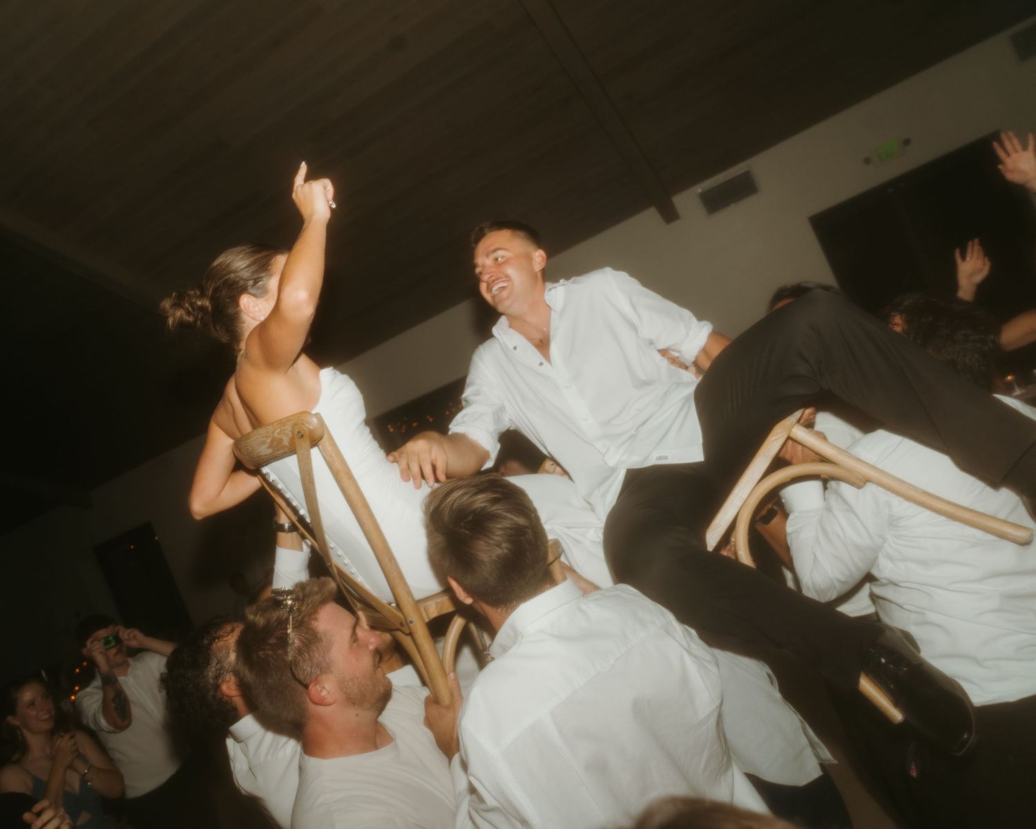 Bride and groom being lifted on chairs during lively wedding hora dance celebration with guests in white shirts.