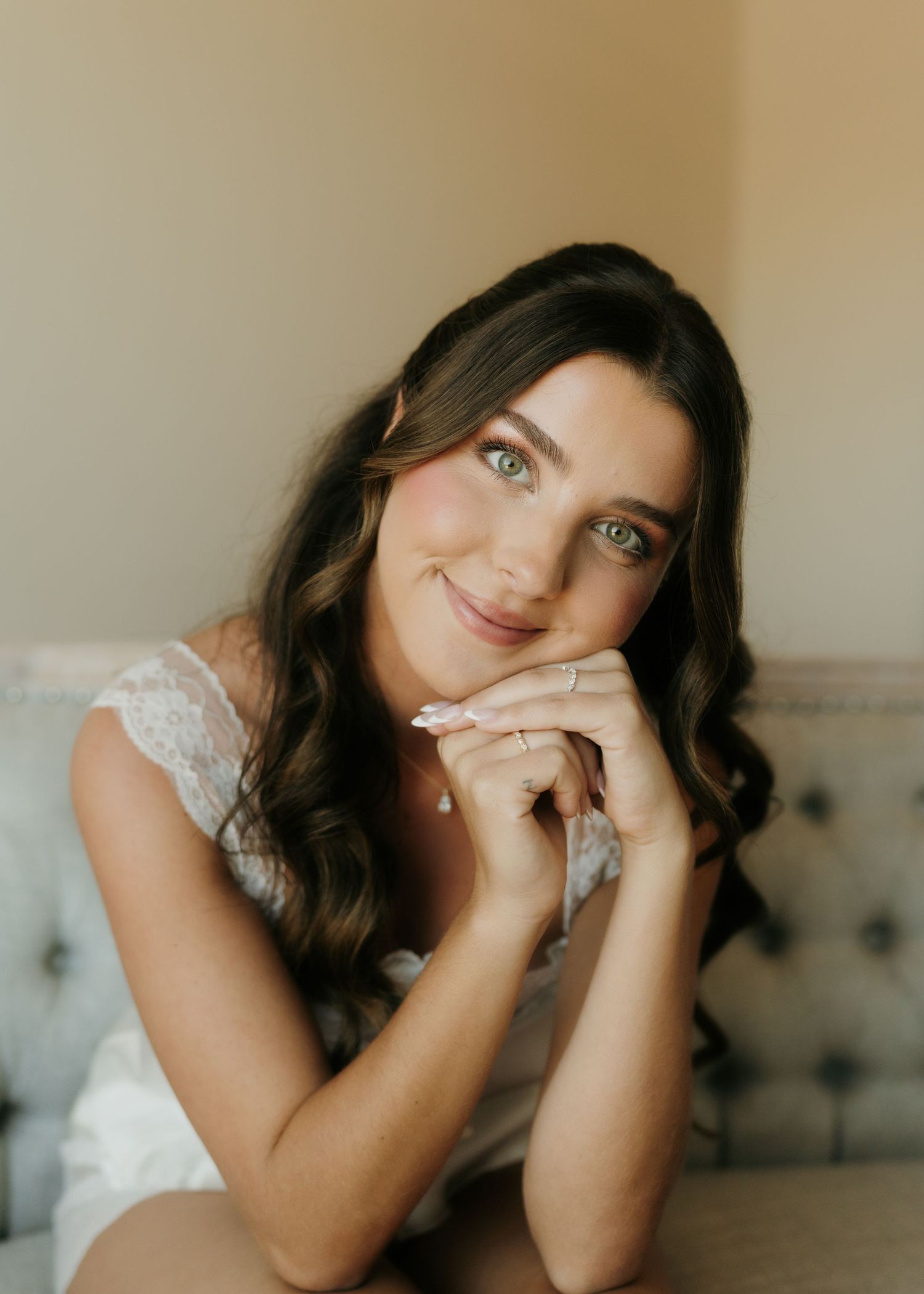 Bride with dark wavy hair and green eyes poses gracefully in white lace, resting chin on hands with a soft smile.