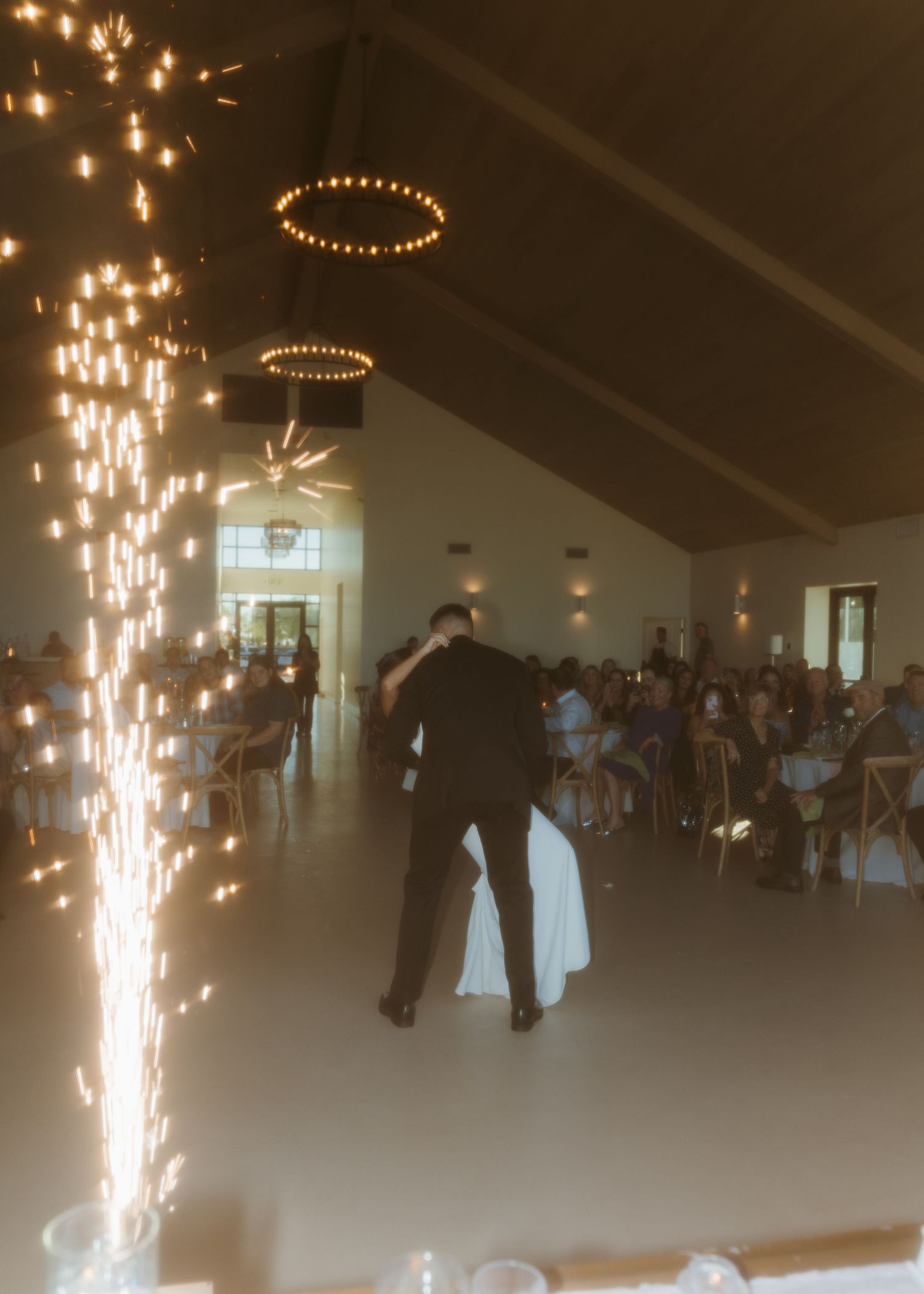 Couple shares first dance at wedding reception with sparkling fountain fireworks and chandelier lighting in elegant venue.