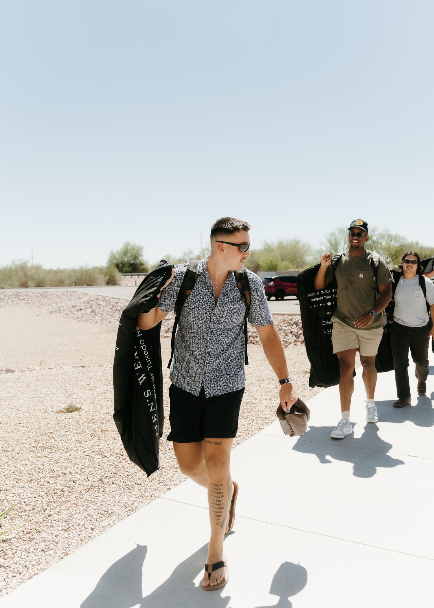Group of people walking outdoors in desert landscape, man in foreground wearing shorts and flip flops carrying jacket.
