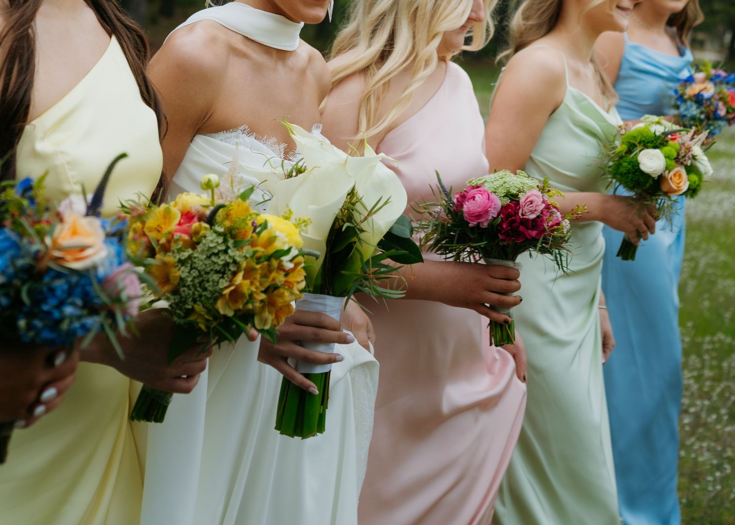 Bridesmaids in pastel dresses holding colorful bouquets with yellow, pink, and blue flowers outdoors.