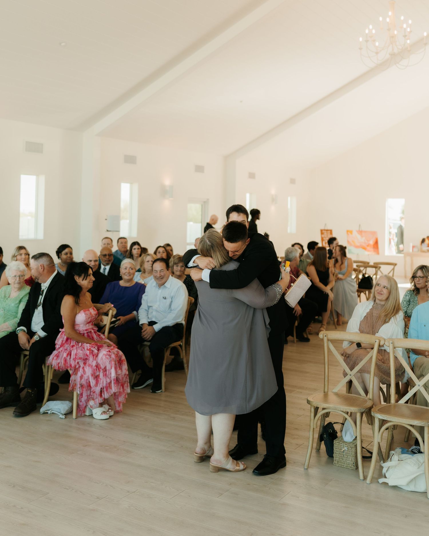 Groom embracing a family member in the aisle of a white wedding venue chapel with seated guests.