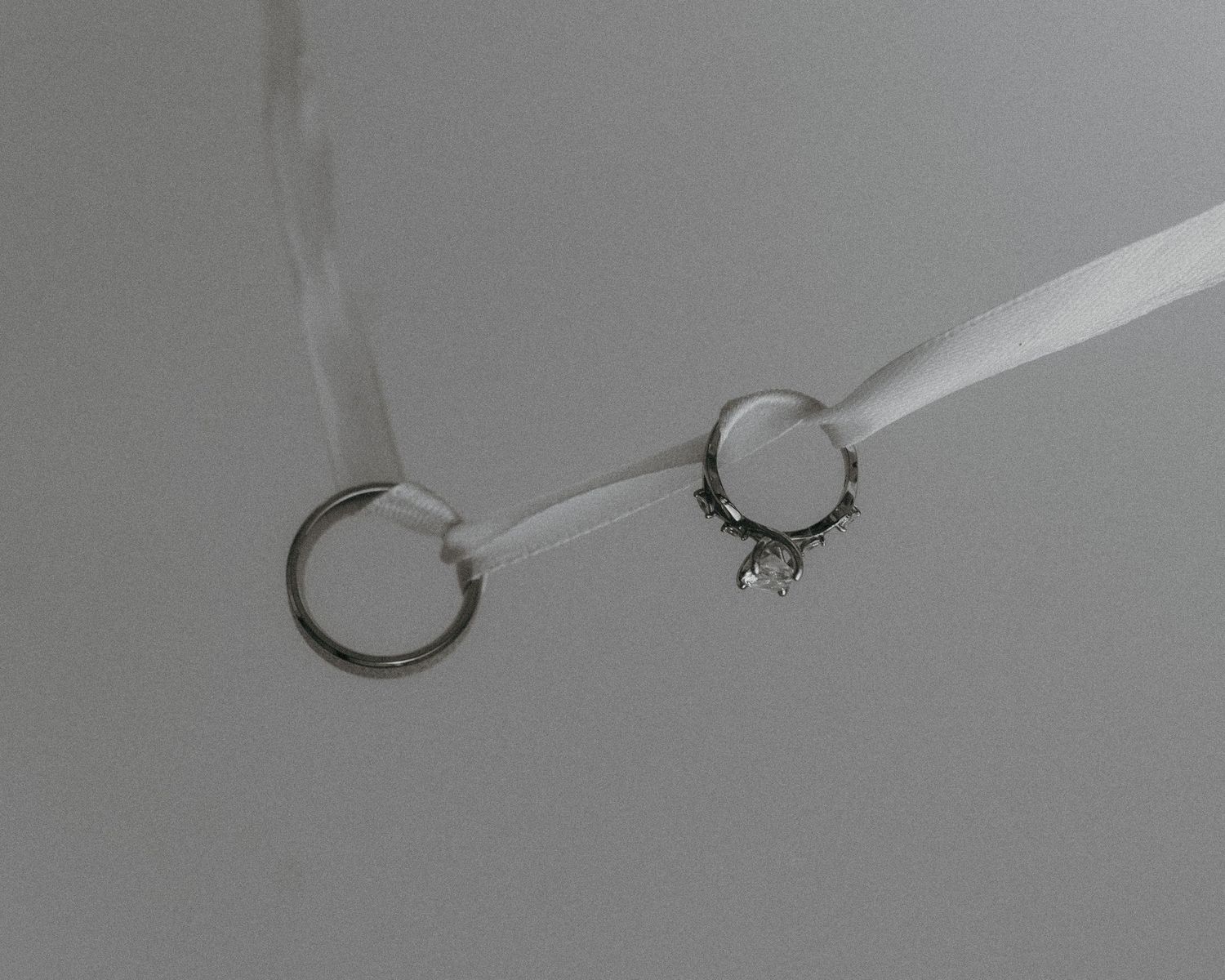 Two silver rings connected by a white ribbon, one plain and one ornate with crystals, on gray background.