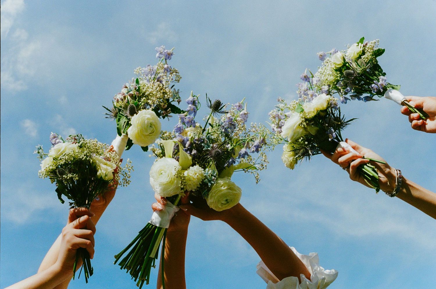Bridesmaids raising colorful wildflower bouquets with white roses and blue flowers against a blue sky.